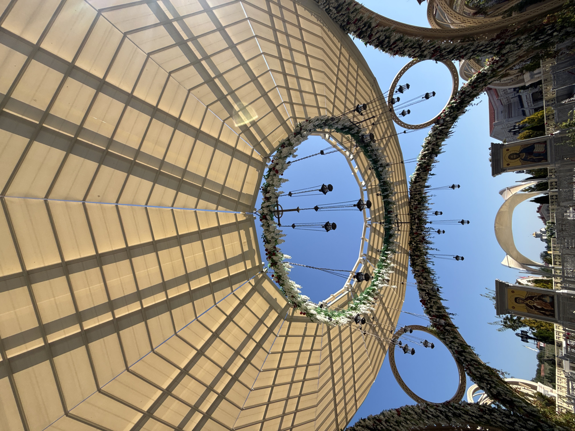 This image shows an outdoor decorative structure with a large, beige, octagonal canopy supported by a metal framework. The canopy is segmented into rectangular panels, casting shadows that create a grid pattern. Suspended from the canopy are several decorative elements, including circular floral arrangements adorned with white and red flowers and greenery. Hanging from these floral rings are multiple small, dark lanterns on chains.
The scene appears to be in an open area with a clear blue sky. In the foreground, there are large, arched structures also decorated with flowers and greenery. Behind these, religious icons are displayed on stands, one showing the Virgin Mary and Child, and another depicting Christ. In the background, buildings with domes and spires, suggestive of a church or religious complex, are visible. The overall impression is of a festively decorated outdoor space, possibly for a religious event or celebration.
The image was taken in Sevastopol, Ukraine, at the Holy Trinity Greek Orthodox Church. This location is known for its ornate architecture and its role as a significant religious and cultural site in the city. The structure in the image is likely part of the church grounds, possibly a temporary installation for an event or a permanent architectural feature designed to enhance the spiritual atmosphere of the location. The presence of religious icons and the architectural style of the background buildings strongly suggest a religious context.