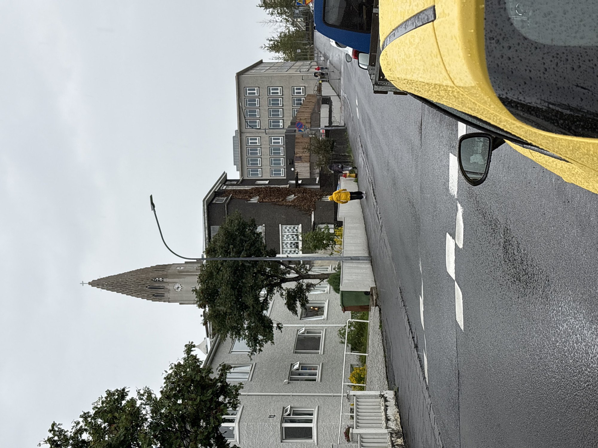 The image captures a street scene in Reykjavik, Iceland, likely on a cloudy or rainy day, given the wet asphalt and water droplets on the yellow car in the foreground. In the background, a tall, gothic-style church tower with a clock face is visible, characteristic of Hallgrímskirkja, Reykjavik's prominent landmark. Several residential and commercial buildings line the street. A person in a bright yellow jacket is walking on the sidewalk, while other figures are further down the street, suggesting moderate pedestrian activity. The road features white pedestrian crossing markings. The overall atmosphere is subdued due to the overcast sky.