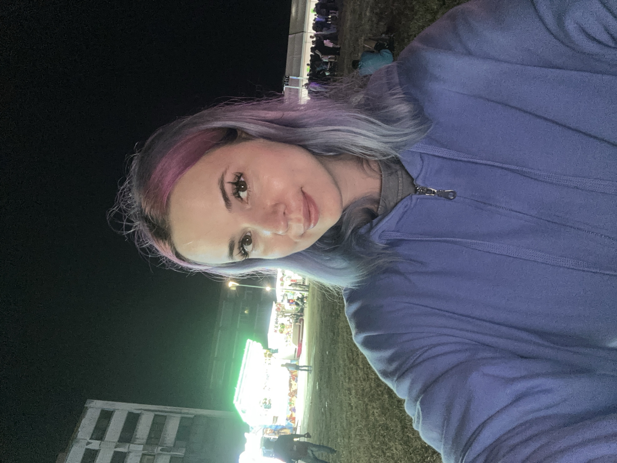 The image is a selfie taken outdoors at night, likely at a fair or festival. A young woman with light skin, dark brown eyes, and dyed purple and pink hair is in the foreground, wearing a blue hooded sweatshirt. Her face is turned towards the camera, and she is smiling. Behind her, there are various structures illuminated with bright lights, possibly booths or carnival rides. Several other people are visible in the background, appearing to be walking or standing in an open area. The ground is covered in grass. To the left of the frame, a tall, multi-story building with a light-colored facade is partially visible against the dark sky. The overall atmosphere suggests a lively event at night.
