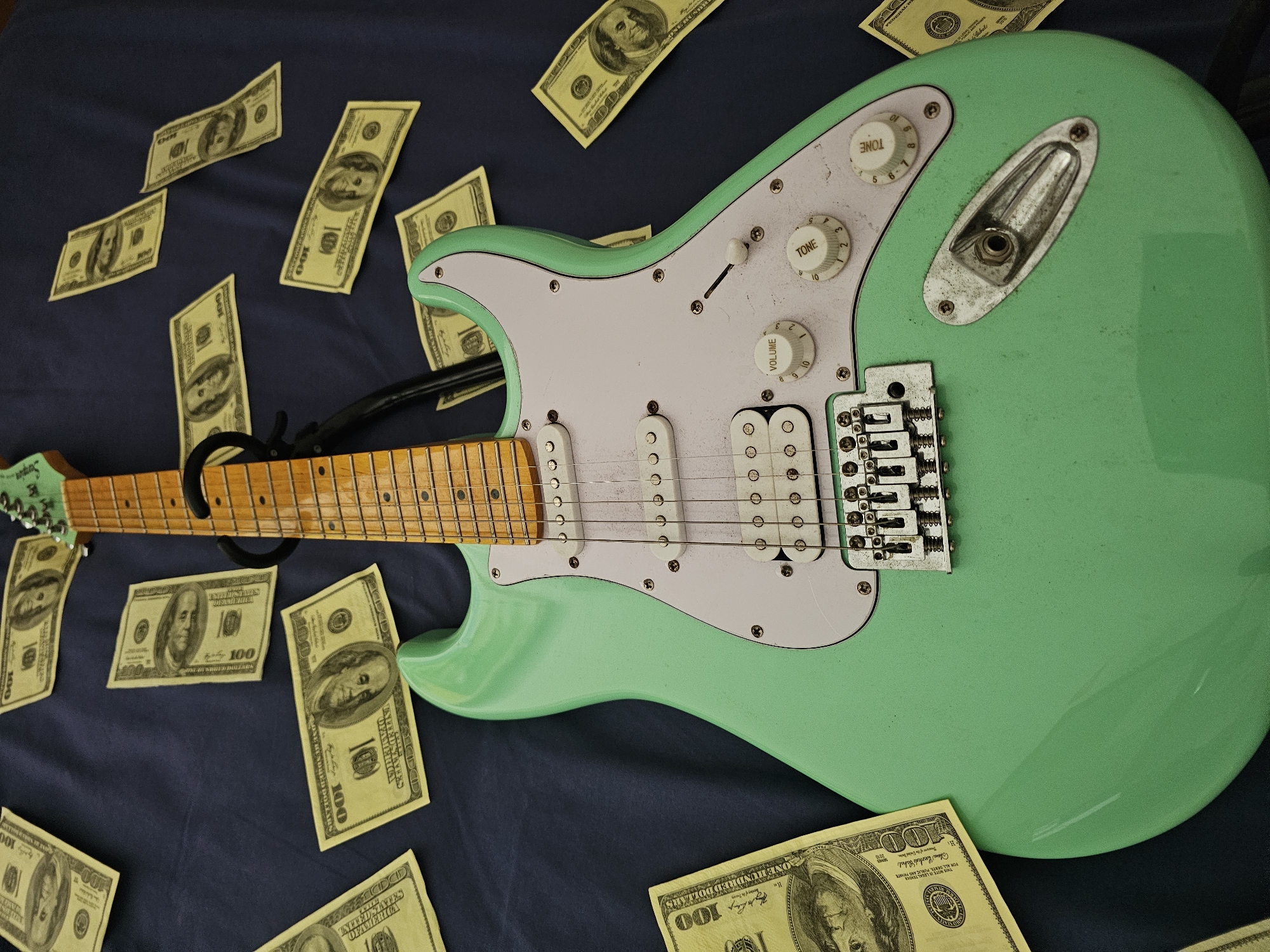 A light green electric guitar lies on a dark blue fabric surface amidst scattered U.S. hundred-dollar bills. The guitar features a maple fretboard and a white pickguard. The scene suggests wealth and music, yet the image provides no indication of being in Denpasar, Indonesia.