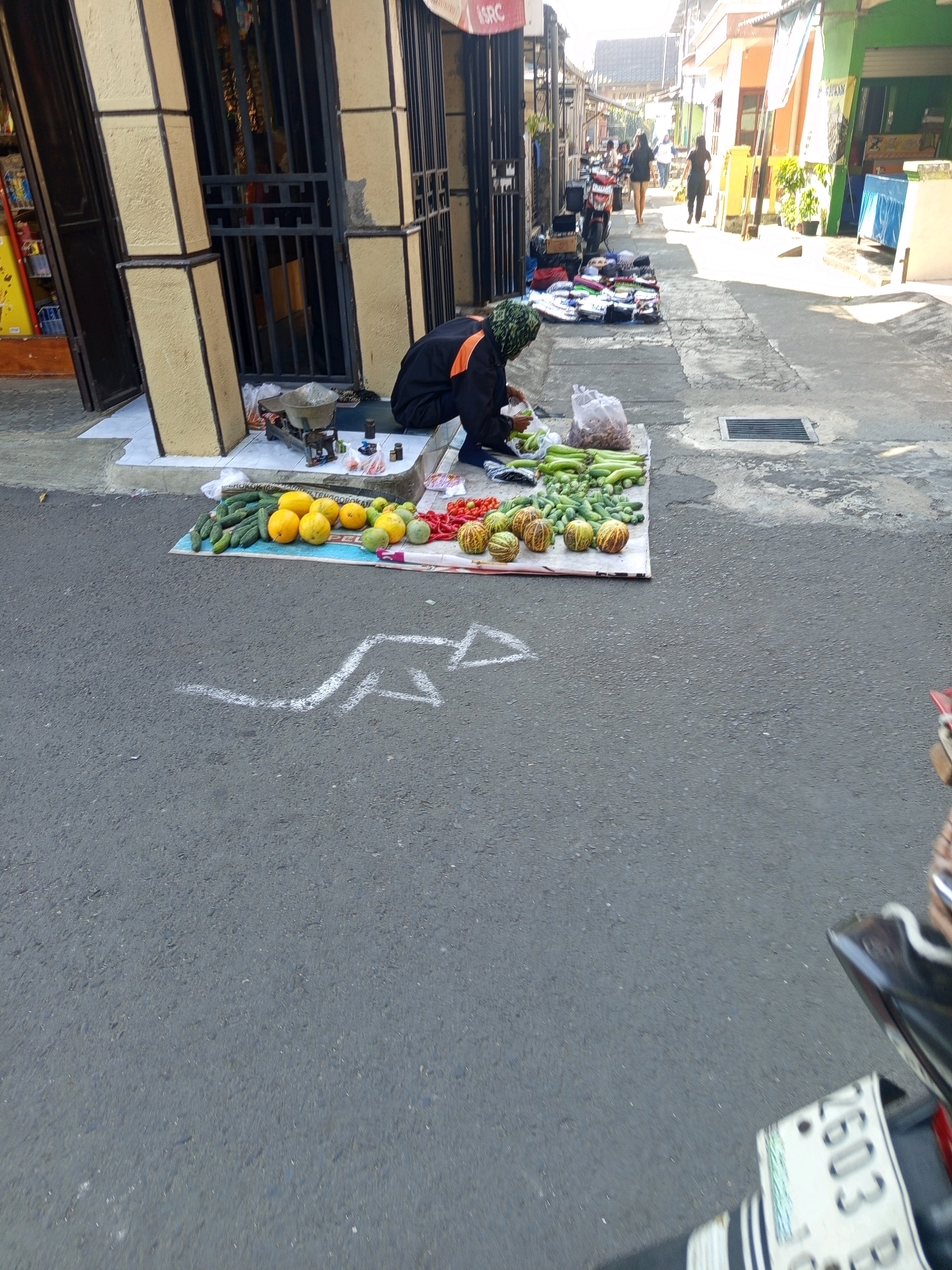 The image captures a street scene in Malang, Indonesia, likely a market area, with a vendor selling fruits and vegetables from a mat laid out on the asphalt. The vendor, dressed in a black jacket and headscarf, is crouched down, attending to the produce. The display includes cucumbers, oranges, melons, and tomatoes, arranged on a patterned mat. In the background, a shop with barred windows is visible, and further down the street, other vendors and shoppers are present, along with parked motorcycles. The sunlight casts distinct shadows, indicating it's daytime. White chalk markings, possibly indicating a lane or boundary, are visible on the road surface in the foreground.