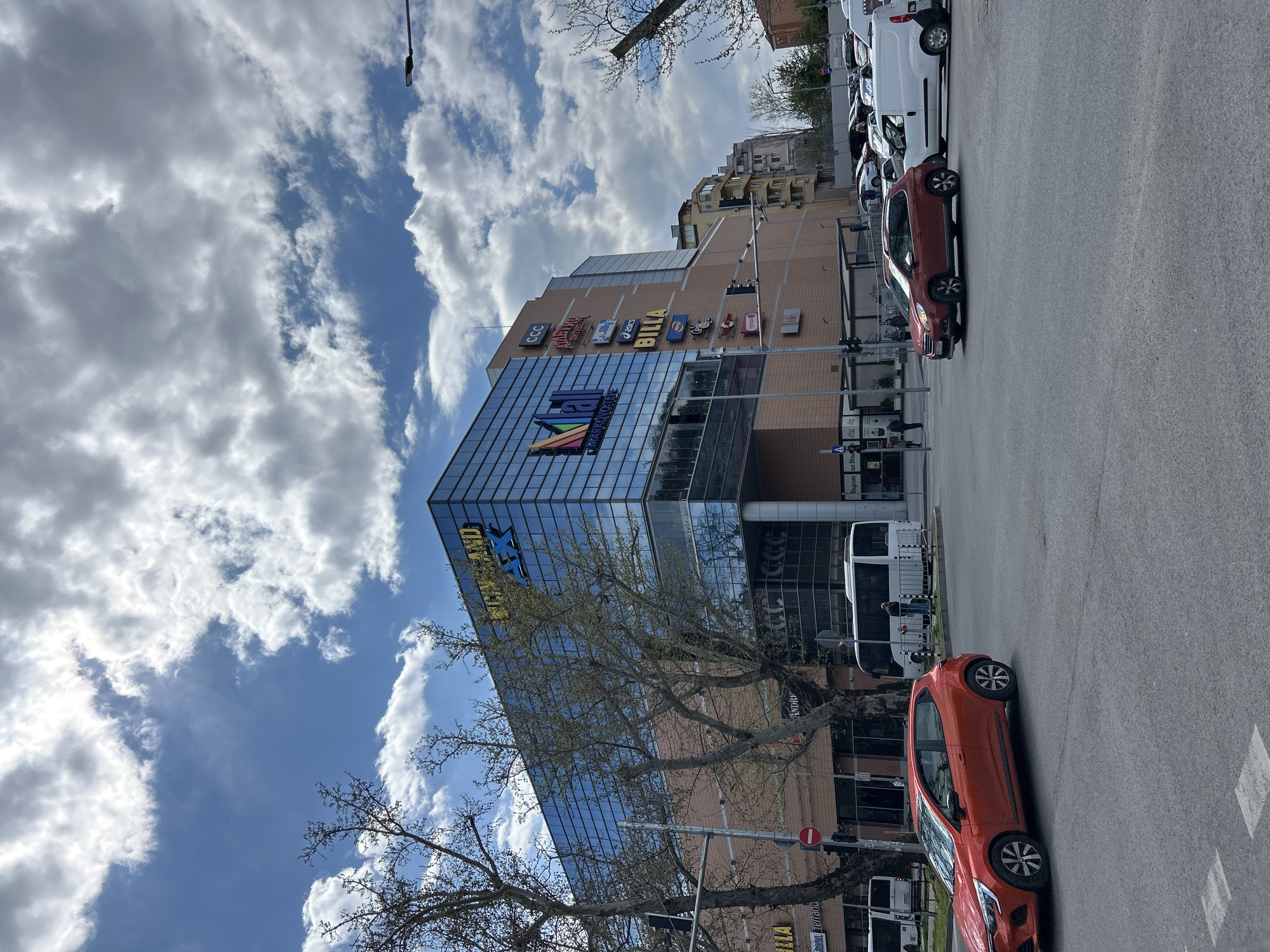 The image shows the Mall Markovo Tepe in Plovdiv, Bulgaria. The multi-story building has a glass facade with "Markovo Tepe" displayed prominently. Various store logos, including Billa, are visible higher up on the building. A partly bare tree stands in front of the building. An orange car is parked on the street in front of the building. A line of cars, including a white van, are parked along the street. The sky is cloudy with patches of blue. Several pedestrians are present, but their actions are indistinct. A traffic light pole and street signage are visible.
