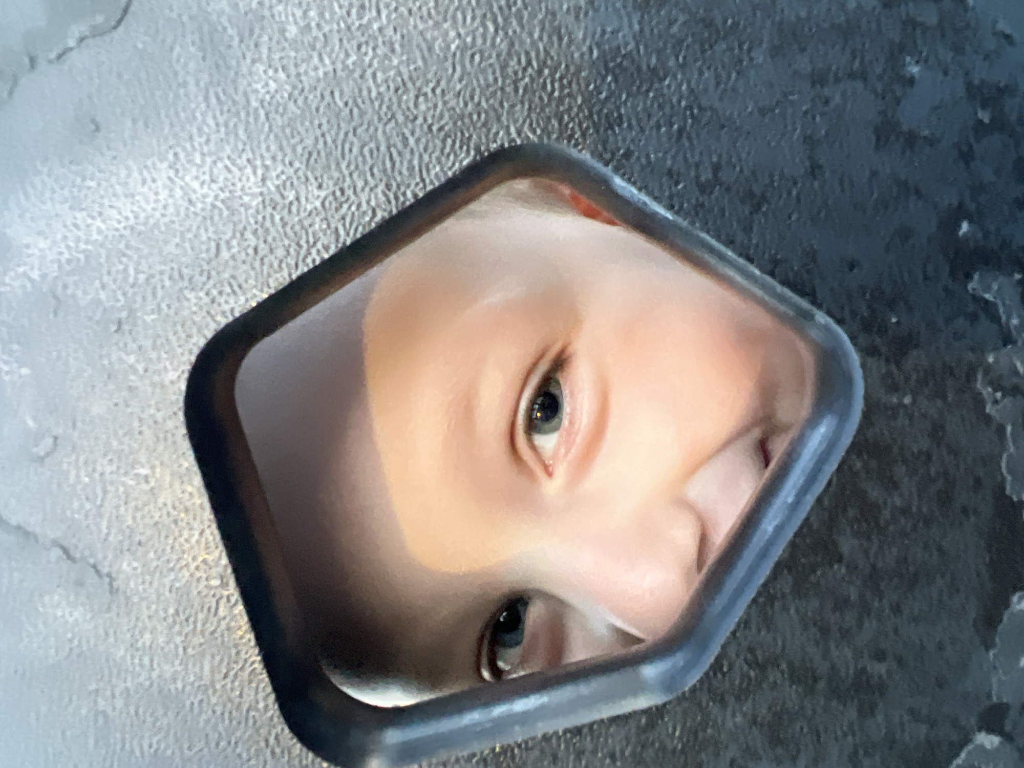 A baby's face is reflected in a small, dark-framed, roughly diamond-shaped mirror. The background is a textured, gray surface, possibly asphalt or concrete. The reflection shows the baby's eyes, nose, and part of their forehead. The surface the mirror sits on is uneven and appears to have slight variations in color. No location specifics are discernable beyond what is presented in the image. No activities or interactions are visible in the frame.