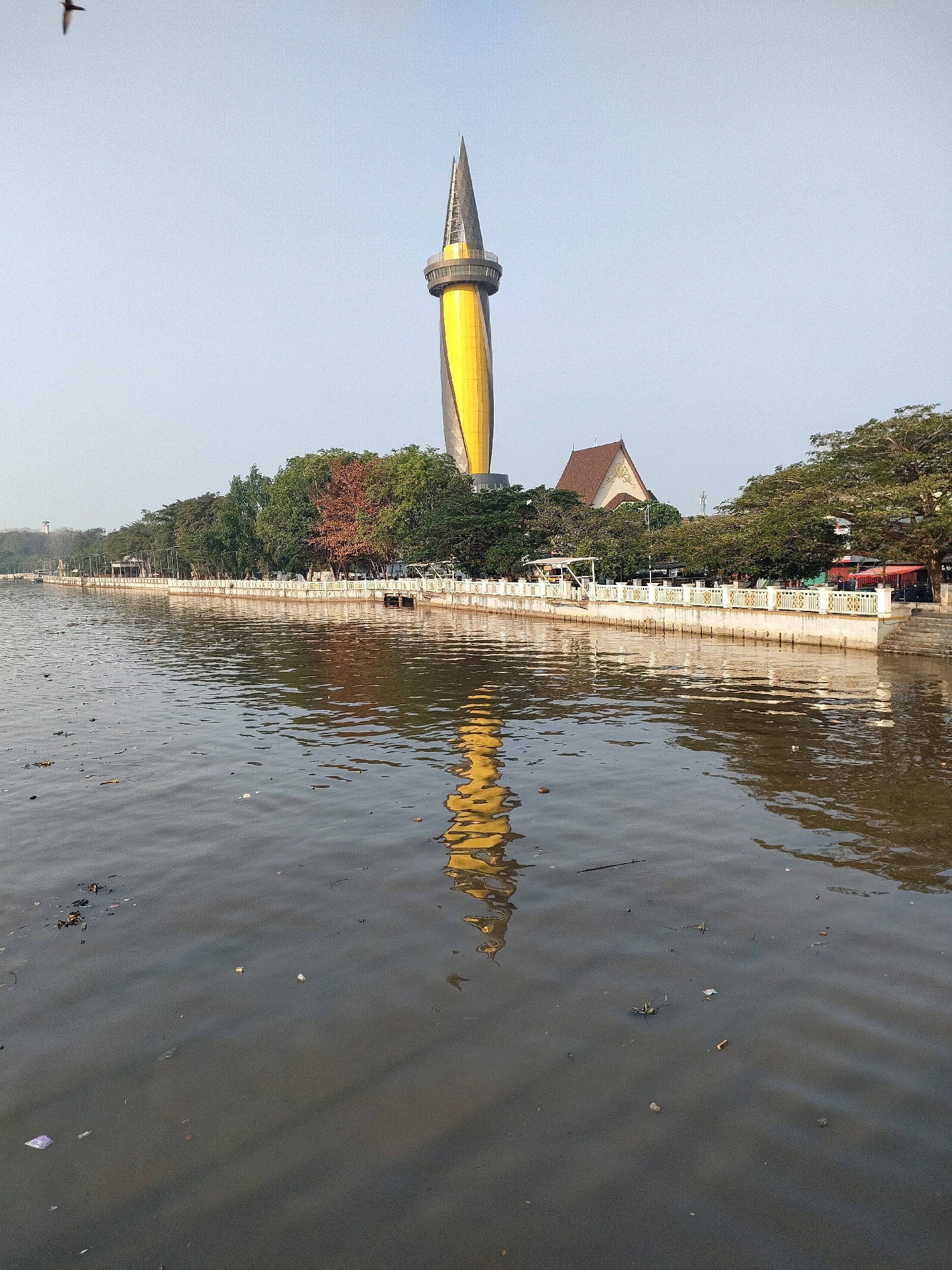 The image shows a canal in Banjarmasin, Indonesia, with a prominent yellow and black spire-like tower reflected in the water. The canal's surface is murky and littered with debris, including plastic bags, wrappers, and other trash. Along the canal's edge, there is a white railing with decorative elements, and behind it, a line of trees. A building with a brown roof and a white facade is visible among the trees, to the right of the tower. The sky is clear and pale blue. A bird is in flight in the upper left portion of the image. The scene depicts a public waterfront area that appears to be in need of cleanup.