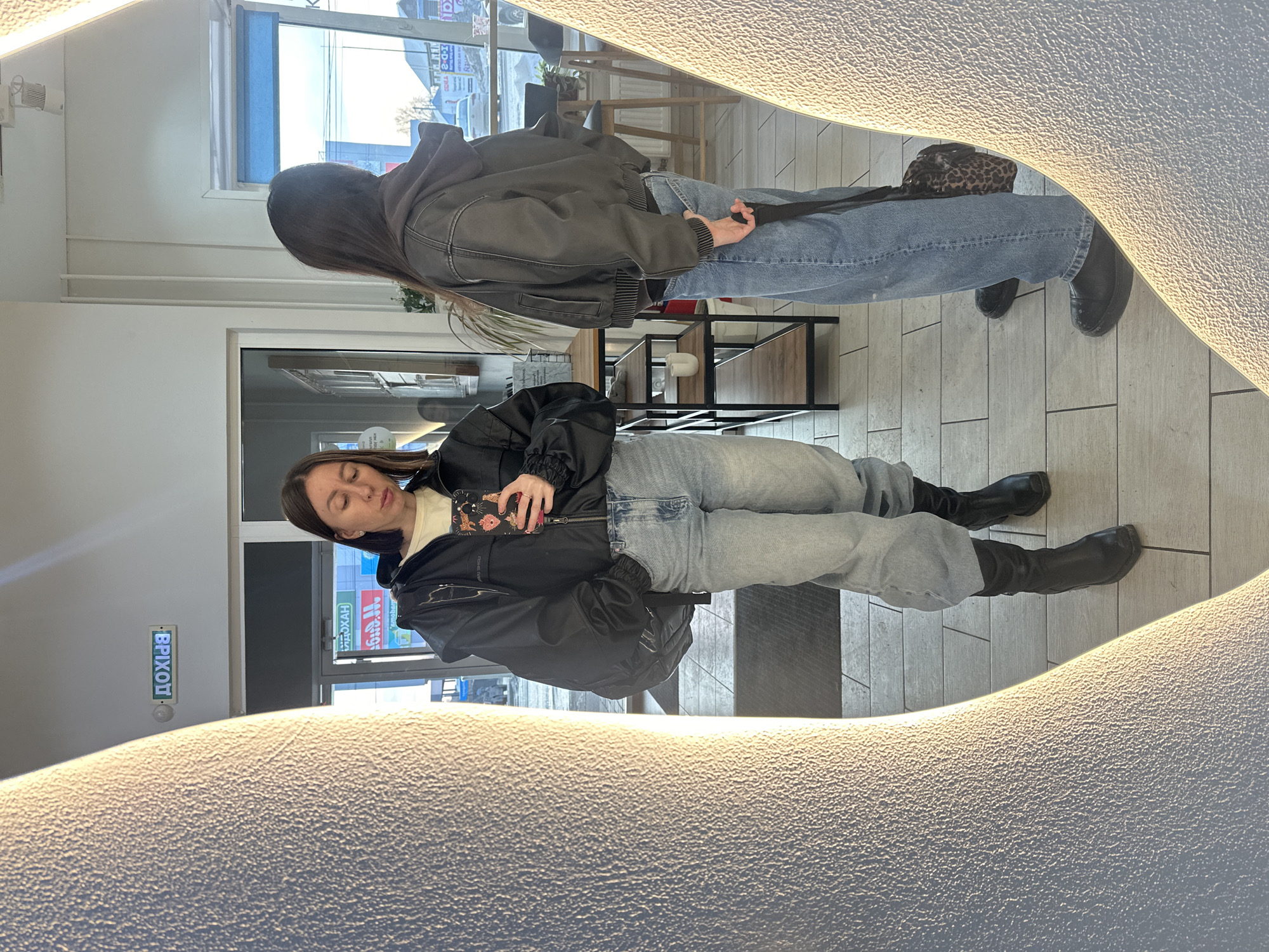 A single female individual is captured in a mirror selfie within an interior space in Votkinsk, Russia. The individual, appearing to be in her twenties or thirties, stands facing the mirror, holding a black smartphone with a colorful patterned case featuring animal-like figures in her right hand, positioned to take a photograph. She wears a black puffer-style jacket over a light-colored long-sleeve shirt, light blue baggy jeans, and black mid-calf boots. A black backpack is slung over her left shoulder, partially obscured by her jacket.
The mirror's reflective surface reveals the individual's back on the right side of the frame, where she is seen wearing a dark grey hooded bomber jacket, the same light blue baggy jeans, and black low-cut shoes. She holds a black strap or handle in her right hand. A leopard-print shoulder bag is visible on her left side.
The background, visible through the mirror and beside the individual, shows a brightly lit indoor area with a light-colored tiled floor. A glass doorway leads to the exterior on the left, displaying an upside-down "ВЫХОД" (VYKHOD - "Exit") sign above the frame and other Cyrillic signs, including "МАГАЗИН" (MAGAZIN - "Shop/Store"), "МОЛОЧНЫЙ" (MOLOCHNYY - "Dairy"), and "2-0-K." The exterior view through the windows reveals a street scene with parked cars, what appears to be bare trees, and buildings under a clear sky, suggesting a cold season.
Inside, behind the reflected individual, a dark metal shelving unit with wooden shelves holds a white cup and other objects. Bar stools and tables are visible further back, implying a commercial setting such as a cafe or shop. Green plants are also present on a shelf and near a window. The mirror itself has a thick, textured, illuminated frame, casting bright light onto the scene.