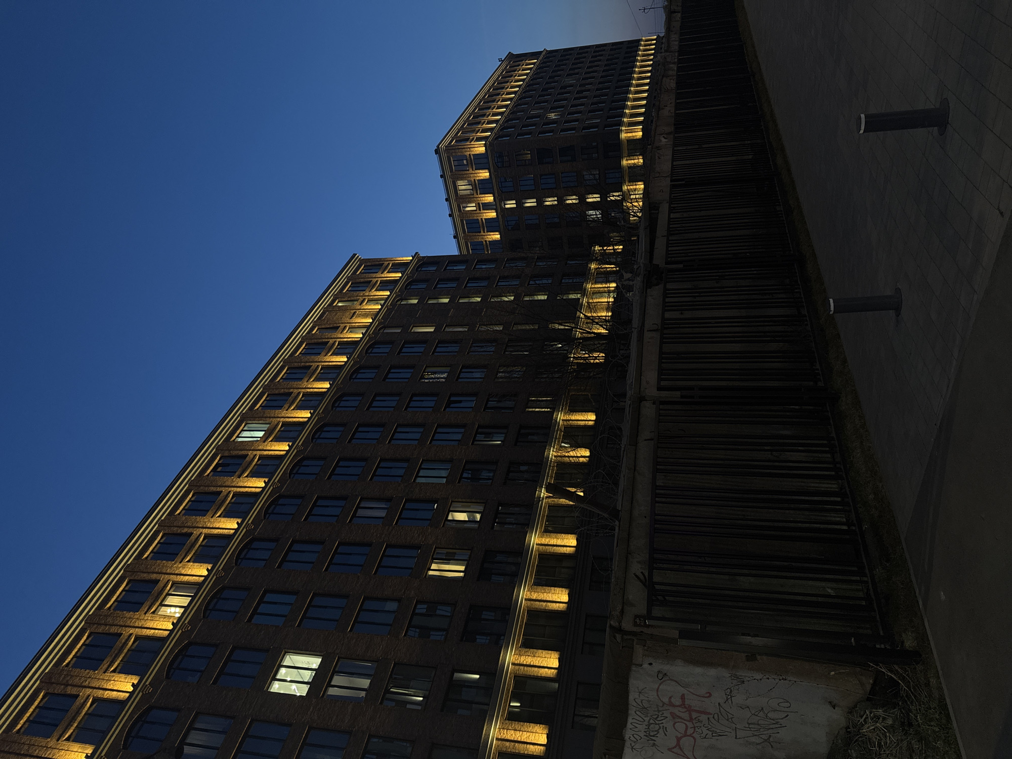 The image shows two tall brick buildings under a deep blue evening sky. The buildings have numerous windows, some illuminated from within. Architectural details on the building's edges are highlighted with yellow light. To the right of the buildings is a dark, decaying structure with graffiti on its surface. Two cylindrical posts are visible on a paved surface adjacent to this structure. The photo is taken from a low angle, looking upwards. The location is not discernable from the image to be Saint Petersburg, Russia.