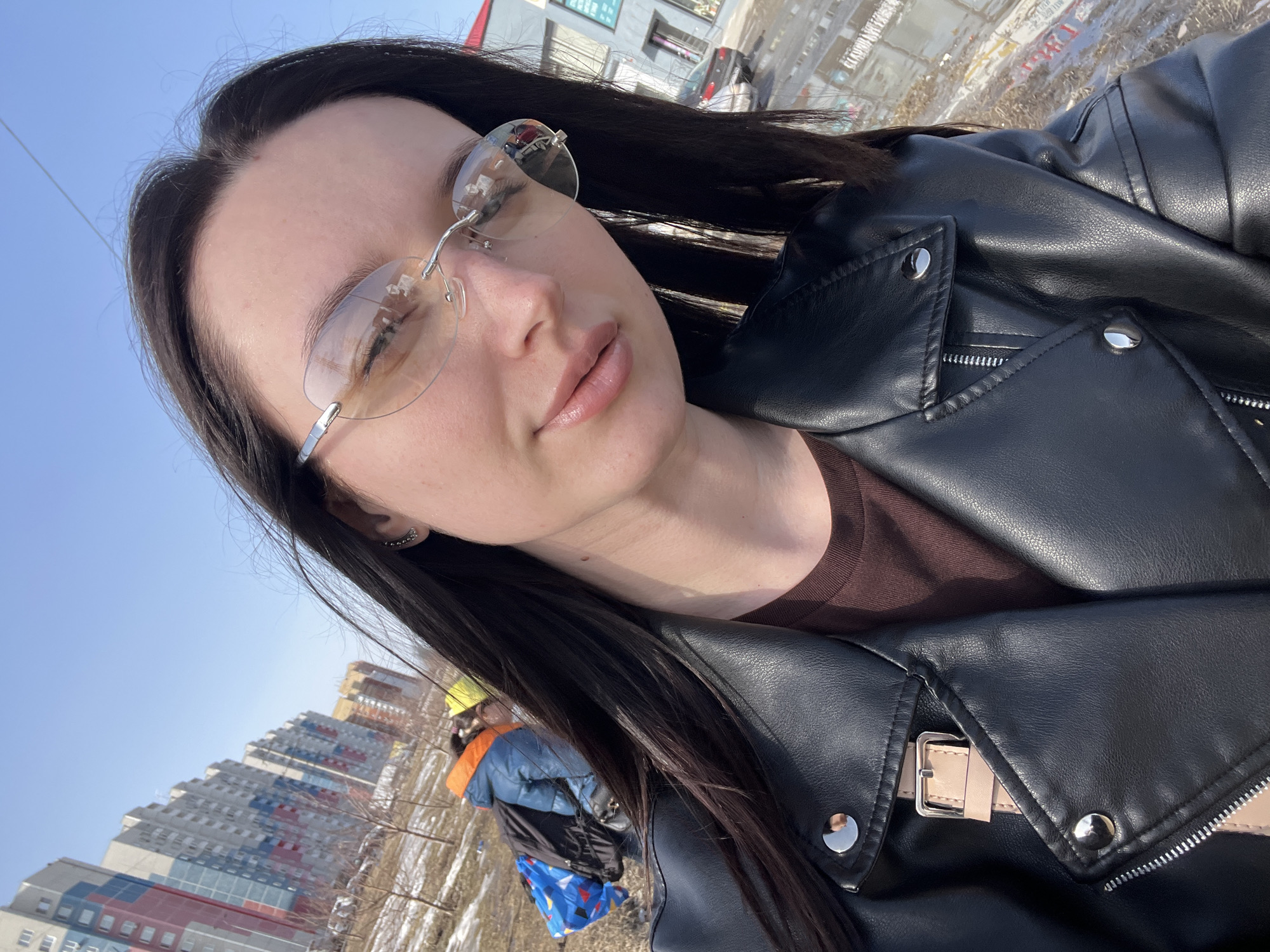 The image presents an outdoor selfie taken by a young adult female in an urban environment in Tyumen, Russia, under clear blue skies. The primary subject is positioned centrally, featuring long dark hair, fair skin, and light-tinted rimless eyeglasses with silver frames. Her expression is neutral with a slight smile, and her lips are natural pink. She wears a black faux-leather motorcycle jacket with prominent silver snaps and zippers, and a beige buckled strap is visible on the jacket's lapel. A brown long-sleeved shirt is worn beneath the jacket.
In the mid-ground, to the left of the subject, two individuals are present on a patch of uneven ground with sparse, bare deciduous vegetation and residual patches of melting snow. One individual, appearing to be a child, wears a blue jacket with an orange element near the neck and a blue backpack featuring colorful geometric patterns, and is leaning forward. A second individual is partially visible behind them, wearing a yellow hat.
The background on the left consists of a row of multi-story residential apartment buildings. The nearest building displays distinct vertical sections of red, white, and blue paneling. Further down the street, other buildings are visible with light grey, white, and some orange or yellow sections. To the right of the subject, a commercial building or storefront with white walls and windows is visible, in front of which a dark-colored sedan is parked on a wet, paved surface that reflects ambient light. The scene is brightly lit by natural sunlight, suggesting a clear day in late winter or early spring.