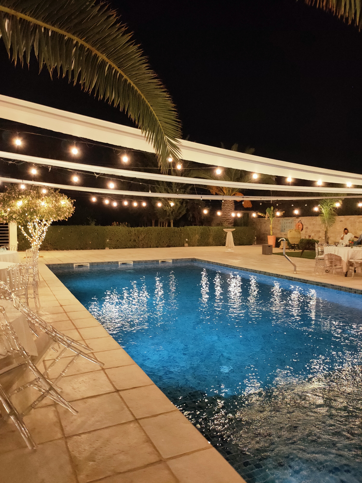 This is a night-time photograph of an outdoor swimming pool area. The pool is illuminated with blue lighting and reflects the numerous string lights that are suspended overhead. To the left of the pool, there are several clear, acrylic chairs arranged near a white-clothed table, suggesting a dining or event setup. A palm frond hangs over the scene from the top left. In the background, there is a manicured hedge, a stone wall, and several potted plants. Two people are visible in the background, seated at a table with white tablecloths. One person is wearing a white shirt. The area appears to be paved with light-colored tiles. The overall scene suggests a festive outdoor gathering or party. This location is in Cebalat, Tunisia.