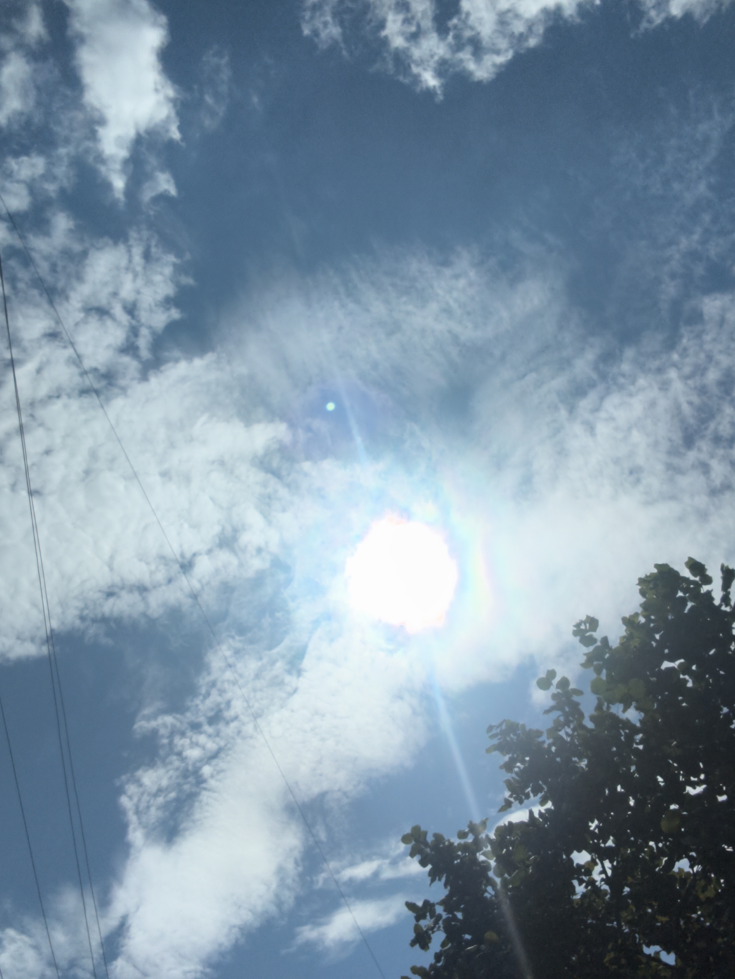 This is a photograph taken outdoors, looking up at a bright sky with scattered clouds. The sun is visible and appears to be at the center of the image, creating a lens flare effect. Several power lines are visible running diagonally across the frame from the left side. In the lower right portion of the image, the dark silhouette of tree branches and leaves can be seen against the sky. The overall impression is of a sunny day with some cloud cover. The location is identified as Chișinău, Moldova.