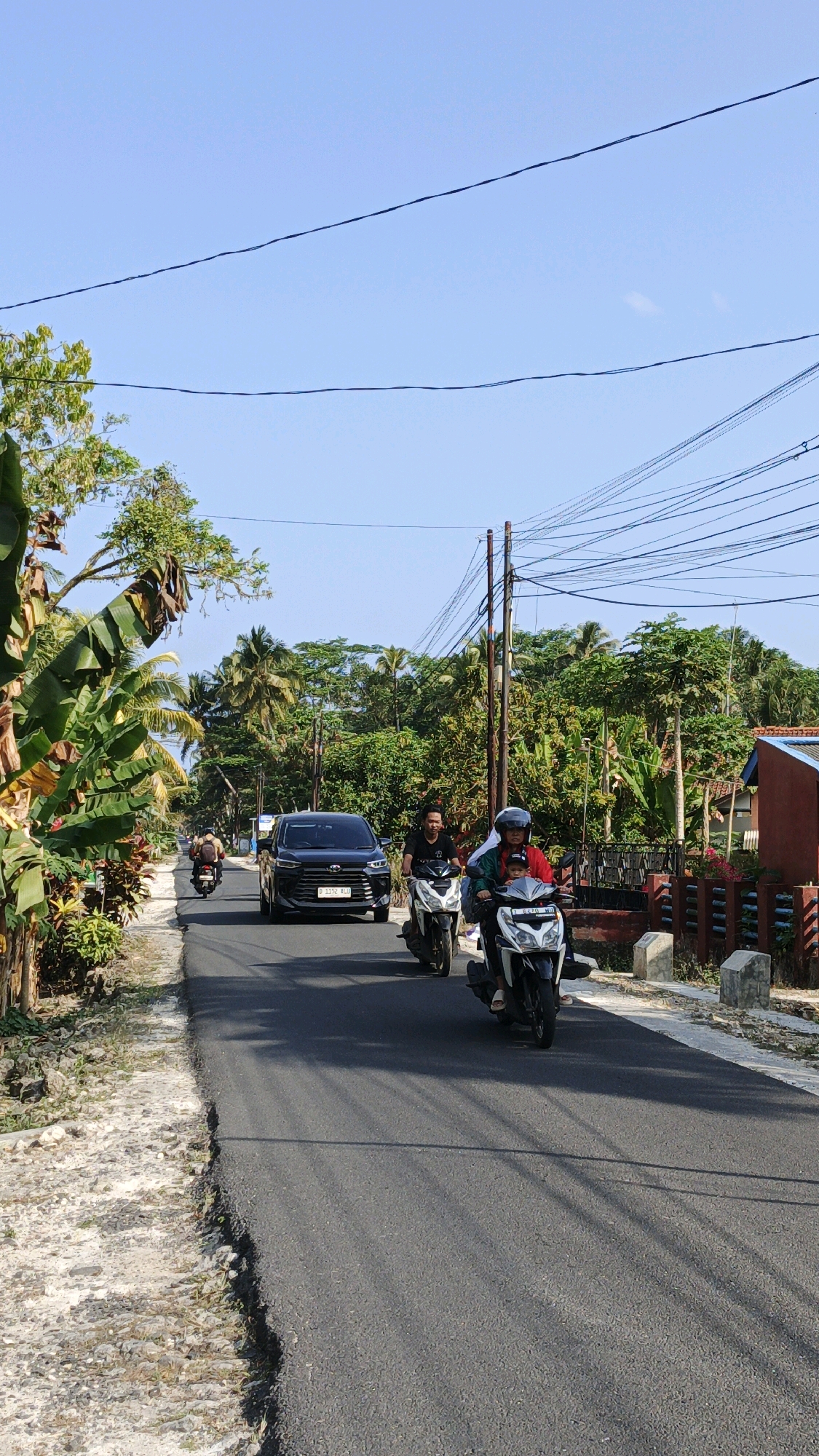 The image depicts a rural road in Cintaratu, Indonesia, under a clear blue sky. On the asphalt road, a black SUV is traveling away from the camera, followed by two motorcycles. The first motorcycle has a rider wearing a black t-shirt and a helmet. The second motorcycle, closer to the foreground, carries two people: the rider in a red shirt and helmet, and a child in the front. To the left of the road, lush green vegetation, including large banana leaves, grows alongside a dirt and gravel shoulder. The right side of the road features a red-colored building with a wooden fence and more trees. Overhead, a complex network of power lines stretches across the sky. The overall scene captures a sunny day on a seemingly quiet Indonesian road.