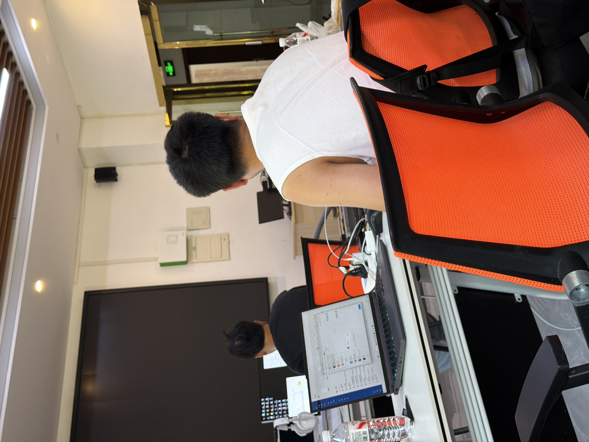 The image shows a bright office environment with two individuals working at desks. In the foreground, a person with short dark hair is seated in an orange and black mesh office chair, facing away from the camera. They are wearing a white sleeveless shirt and have their right arm resting on the armrest of the chair. In front of them is a dark laptop with an open screen displaying a user interface with text and icons. To the left of the laptop, a clear plastic water bottle with a red label is visible. In the mid-ground, another person, also facing away from the camera, is seated at a desk with their back to the viewer. They are wearing a black t-shirt and appear to be working on a computer. Other office equipment, including monitors, headphones, and cables, are also present on the desks. The background shows a wall with some framed documents and a large black screen. The ceiling has fluorescent lighting and wooden accents. A door with a golden frame is visible on the right side of the image, with a green exit sign above it. The overall impression is a typical office setting in a modern building. This scene is located in the city of Jiangjin, China.