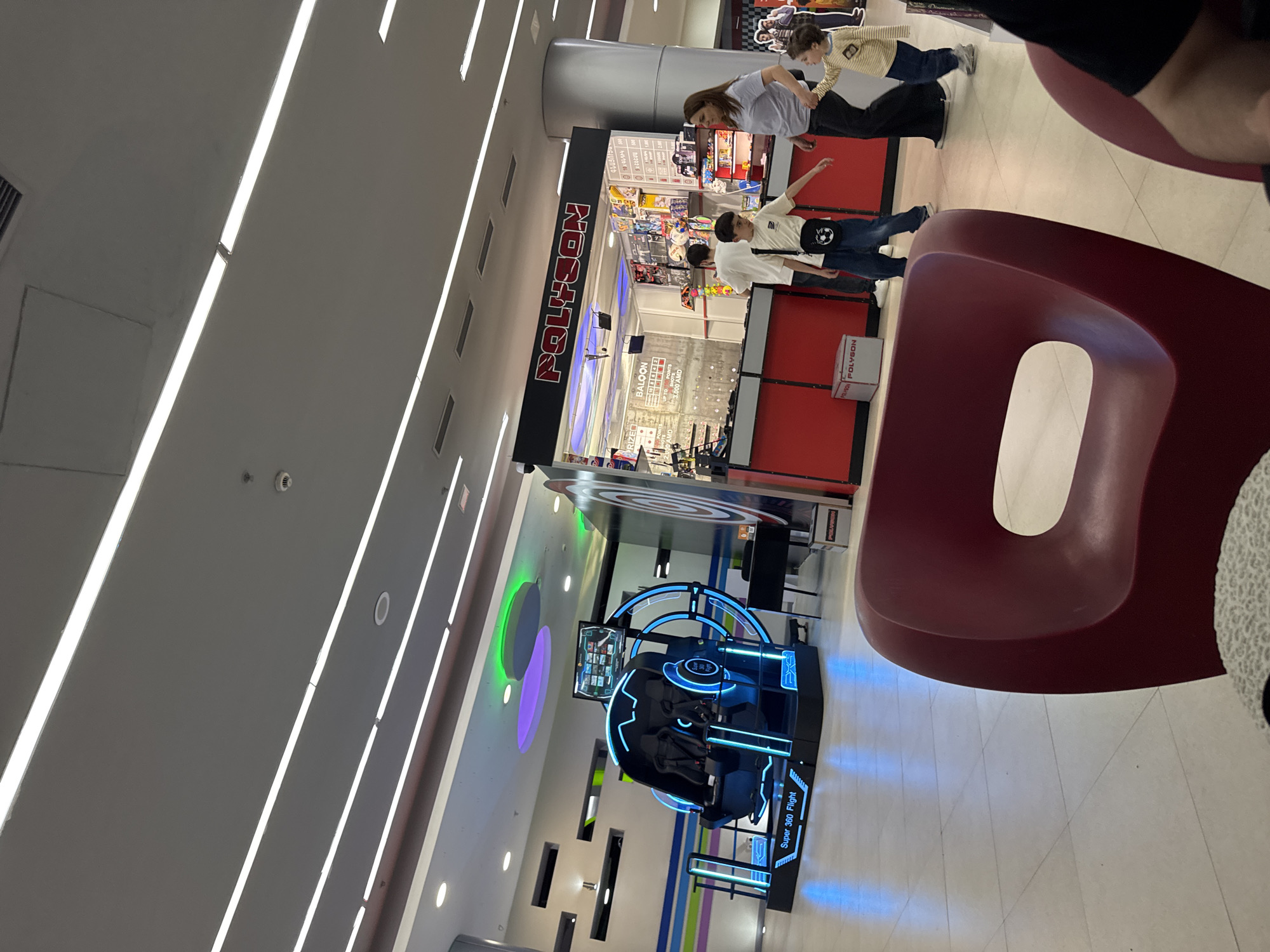 This image depicts an indoor entertainment area, likely a shopping mall arcade, characterized by bright lighting and modern furnishings. The setting is identified by visible text and the provided context as being in Yerevan, Armenia.
In the midground, a prominent "POLYSON" arcade store is visible, featuring a bold black and red sign. Inside the store, various games and displays can be seen, with a sign partially visible that reads "BALOON" and another indicating a price of "3000 AMD," confirming the Armenian location context. A "POLYSON" branded box is also on the floor near the entrance.
Several people are present in the scene. A woman in a grey t-shirt and black pants walks past the Polyson store, holding hands with a child dressed in a striped long-sleeve shirt and blue pants. Another individual, wearing a white t-shirt, blue jeans, and a black shoulder bag with a white soccer ball design, appears to be interacting or gesturing towards the store. In the foreground on the right, a person in dark clothing is partially visible, seemingly seated on a distinctive red, modern-shaped bench or chair.
To the left of the Polyson store, a "Super 360 Flight" virtual reality simulator with glowing blue neon lights is a focal point, indicating interactive gaming opportunities. The overall environment features a light-colored tiled floor and a ceiling equipped with multiple long, linear fluorescent lights and some recessed lighting fixtures, along with a visible smoke detector. The atmosphere is casual and engaging, typical of a public leisure space or entertainment complex.