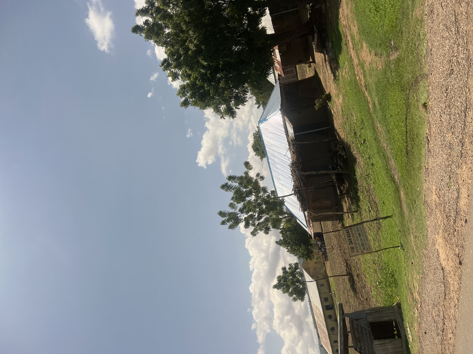 The image depicts a rural outdoor scene within the Balanga Local Government Area, Gombe State, Nigeria, as confirmed by a prominent sign. The foreground consists of a reddish-brown gravel and dirt path, transitioning into areas of green grass and compacted earth paths. On the far left, a small, rustic wooden structure resembling a shack, with an open entrance, is present.
A wooden signpost stands in the central midground. Its text reads "YOBI DAN-MAMA," "SHOPPING COMPLEX FASHION DESIGN," "NURSERY LCHA YARJAL," "BALANGA LGA GOMBE STATE," and "WASH AREA COMPLEX BY GOVT SCHOLAR," indicating the functions of the area.
Multiple buildings are visible. To the right of the sign, a compact, single-story building features a shiny, corrugated metal roof and dark, textured walls, likely constructed from natural materials such as mud or wattle and thatch. Further right, a large, dense tree partially obscures another building with a rusty corrugated metal roof. In the left midground, two larger, rectangular buildings with light-colored walls and corrugated metal roofs stand on a gentle, grassy incline.
The environment includes several trees with green foliage. A large tree dominates the far right, while multiple smaller, lighter-foliaged trees are distributed across the midground. A small cluster of three to five individuals is visible near a tree between the central and left-side buildings; their activities are not discernible. The background on the left horizon reveals distant, hazy hills beneath a sky with scattered white cumulus clouds. The scene is brightly illuminated by direct sunlight.