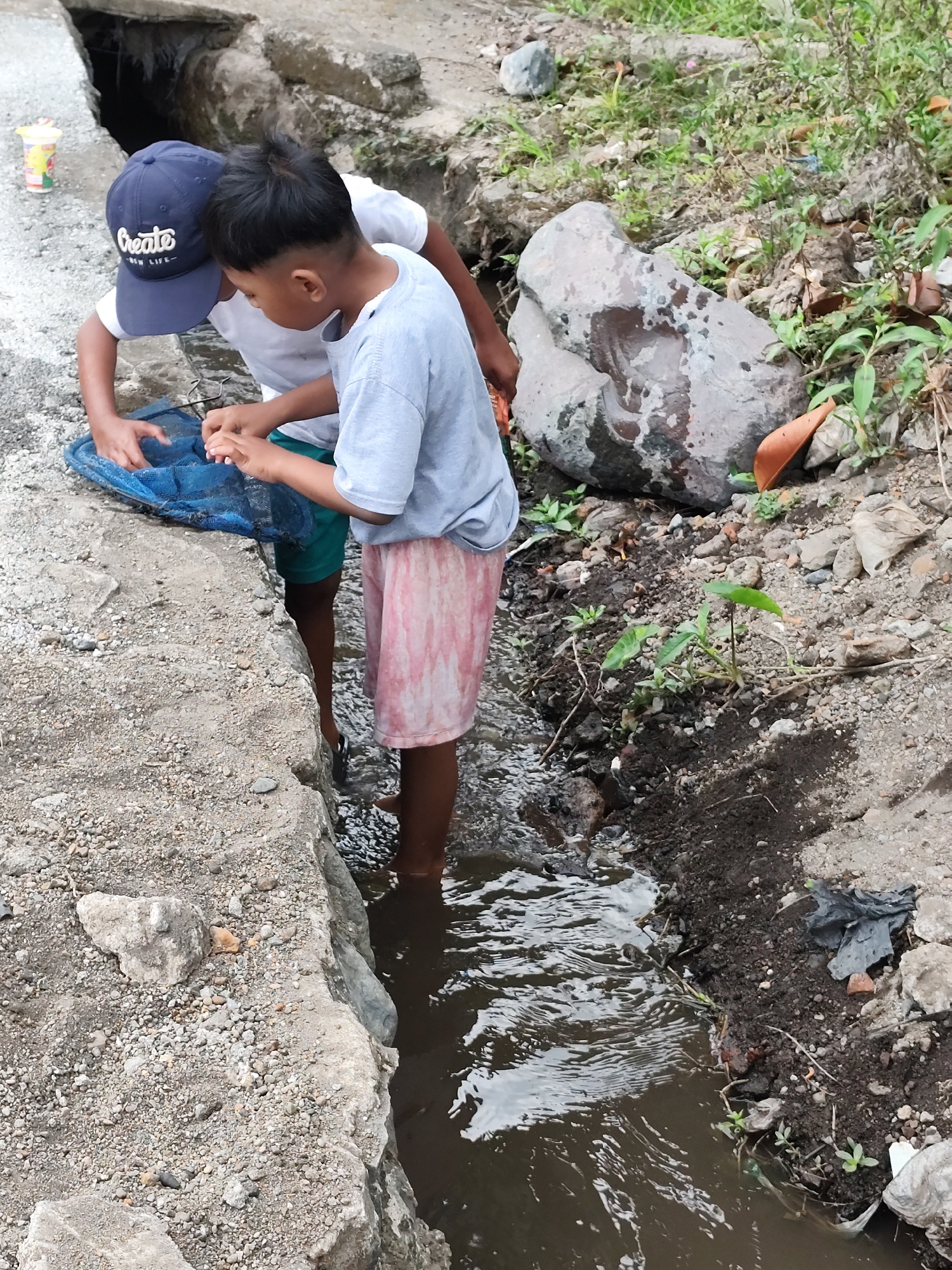 Two young boys are engaged in fishing in a shallow, murky stream at the edge of what appears to be an urban or semi-urban area in Jambewangi, Indonesia. One boy, wearing a blue baseball cap with "Create" and "New Life" printed on it, leans over a blue fishing net, his hands submerged in the water as if attempting to catch something. The other boy, wearing a light blue t-shirt and a pink and white patterned skirt, stands next to him, also with his hands near the net. Their legs are immersed in the stream, which flows through a concrete channel. The surrounding environment consists of rough, rocky terrain with sparse vegetation and some debris, including a discarded plastic cup and plastic bags. The scene suggests a casual, everyday activity taking place outdoors in a somewhat neglected area.