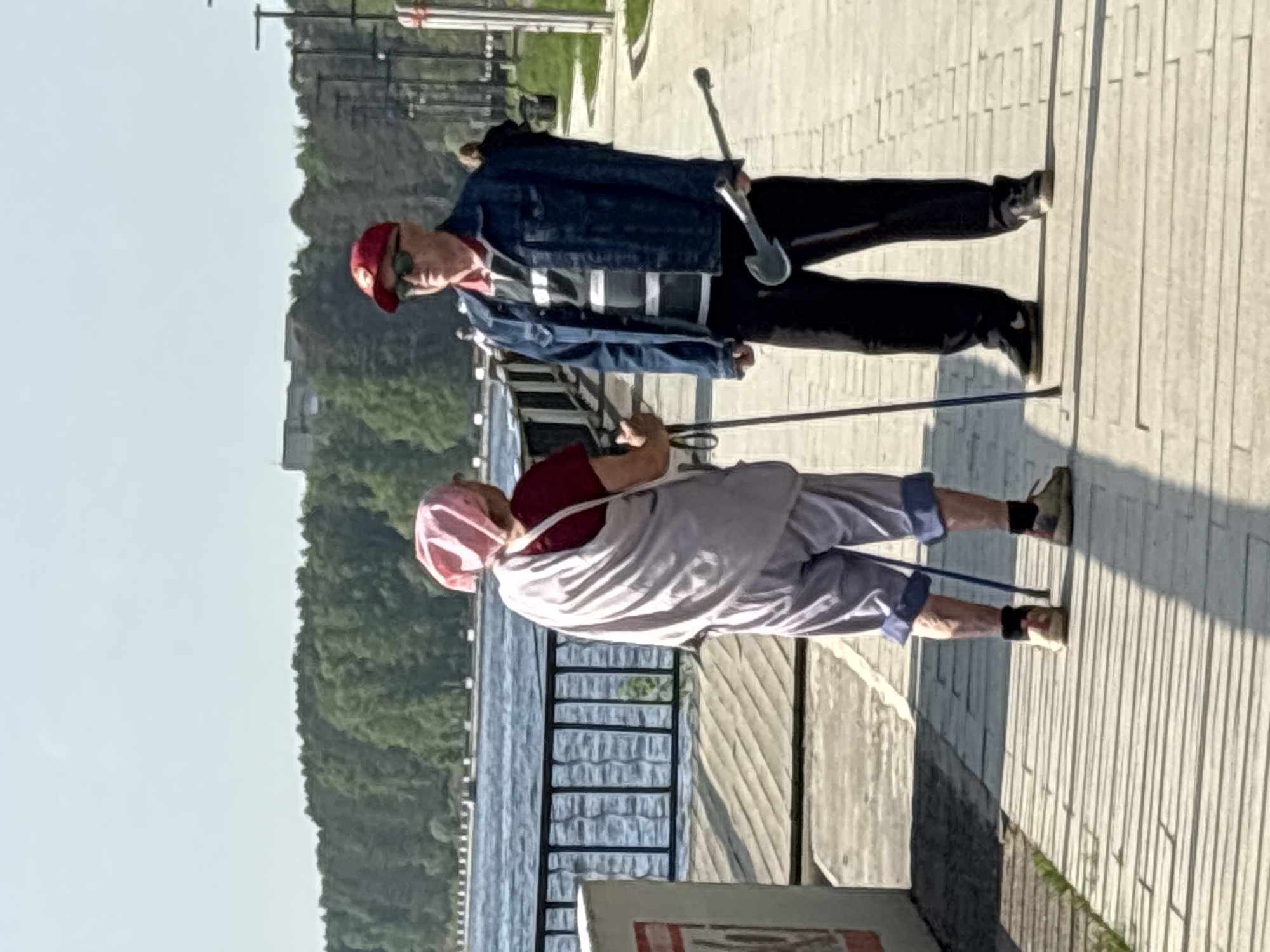 Two elderly people are walking along a waterfront walkway. One person, wearing a red cap, sunglasses, a denim jacket over a striped shirt, and black pants, is using a metal walking stick with a shovel-like end. The other person is wearing a pink head covering, a red shirt under a white shawl, and light purple pants with dark blue cuffs. They are using walking poles. They are walking alongside a metal railing. In the background, there is a body of water, trees, and buildings. The walkway is constructed of long, light-colored planks. The photo likely depicts a scene in the city of Chaikovsky, Russia due to the presence of the waterfront walkway and elderly individuals.