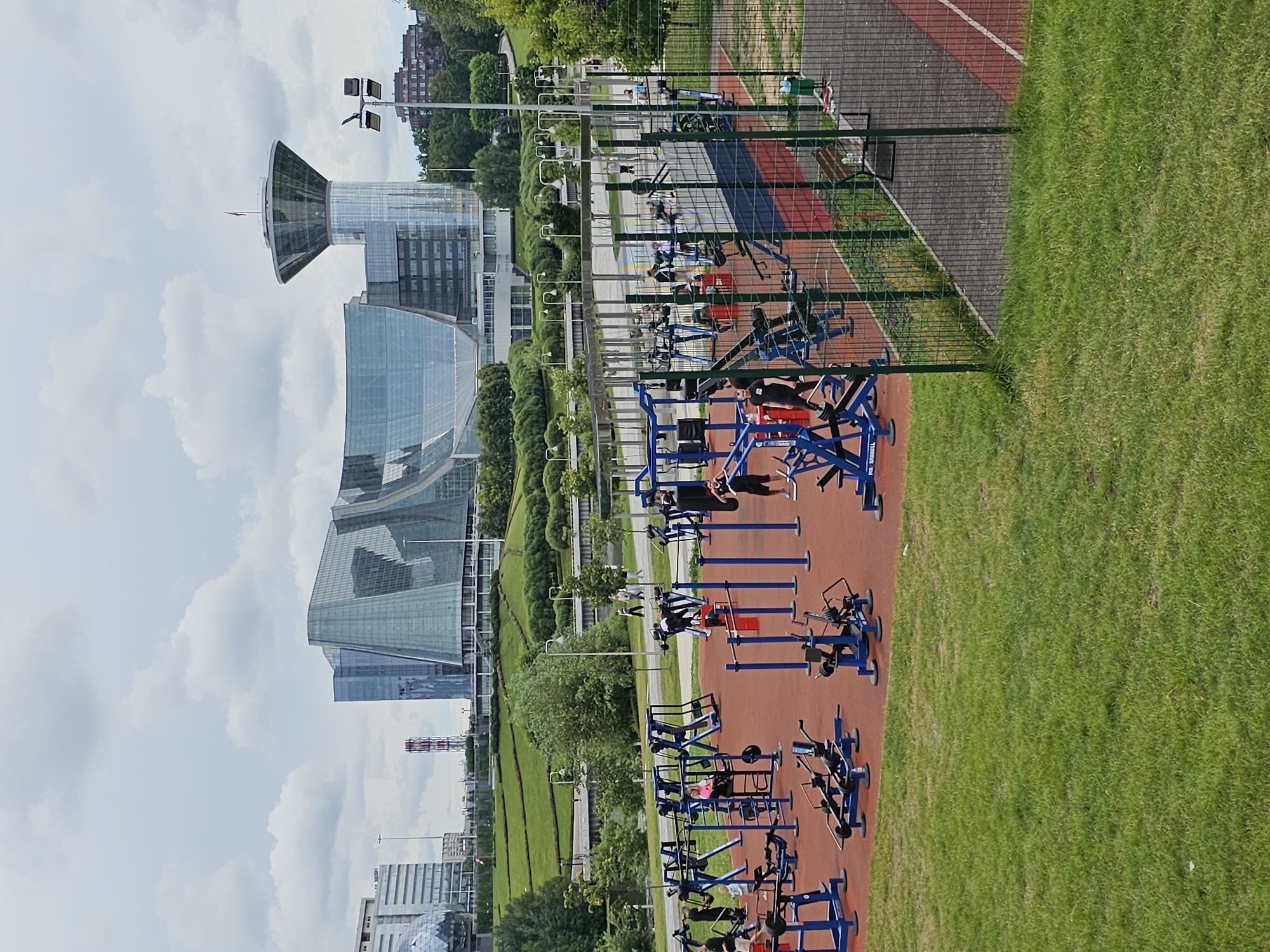 The image depicts an outdoor public gym area located in Khimki, Russia, with modern glass-facade buildings in the background. The gym features numerous blue and red exercise machines arranged on a red rubberized surface. Several individuals are visible actively using the equipment, engaging in various strength training exercises. Some people are standing or sitting on the machines, while others are performing repetitions. The surrounding environment includes a well-maintained lawn, a paved pathway, and a fenced area with a running track and a bench. The sky is overcast with scattered clouds, suggesting a temperate day. The architectural style of the background buildings is contemporary, characterized by sleek lines and extensive use of glass.