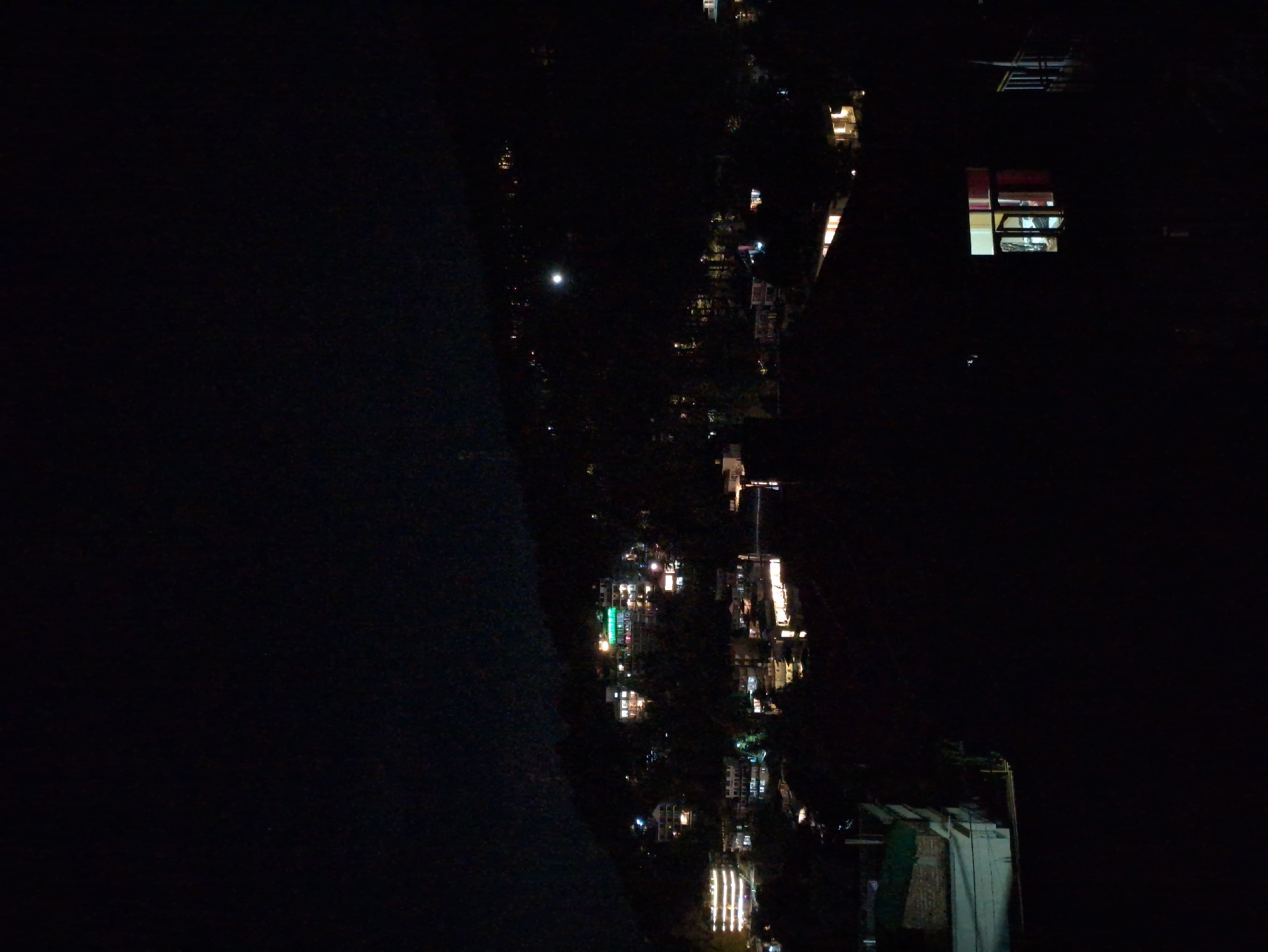 The image captures a nighttime view of a hillside city, with lights from buildings illuminating the darkness. In the foreground, a dark shadow obscures much of the scene, but a window with light emanating from within is visible. Further back, numerous buildings are scattered across the slopes, their windows and architectural features glowing against the night sky. Some buildings appear to be hotels or larger structures, with multiple illuminated floors and balconies. The overall impression is of a settled, populated area with ongoing activity, despite the late hour.