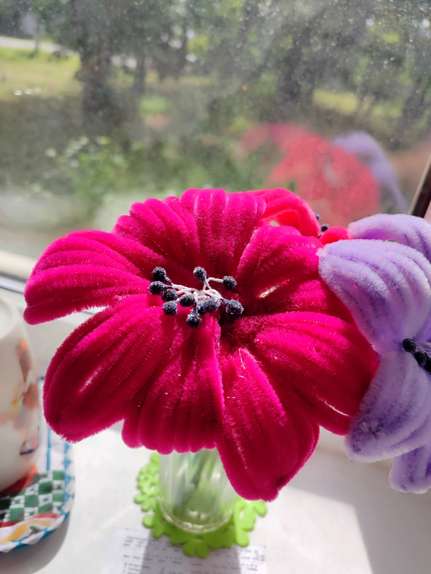 A close-up, eye-level, vertical shot features two artificial flowers, one bright pink and the other lavender, positioned on a sunlit windowsill. The pink flower, the primary subject, is prominently displayed with its petals unfurled, showcasing a velvety texture and a dark stamen. To its right, the lavender flower is partially visible, its petals also textured and vibrant. Both flowers are set against a blurred background of green foliage and trees, suggesting an outdoor environment viewed through the window. The windowsill itself appears to be white, with a clear glass vase holding the flowers and a green, flower-shaped coaster underneath it. A small stack of papers is also visible on the windowsill. To the left of the flowers, a portion of a white mug with a floral pattern peeks into the frame, resting on a colorful woven mat with a checkered pattern. The lighting suggests it is daytime, with sunlight casting shadows and highlights on the scene.