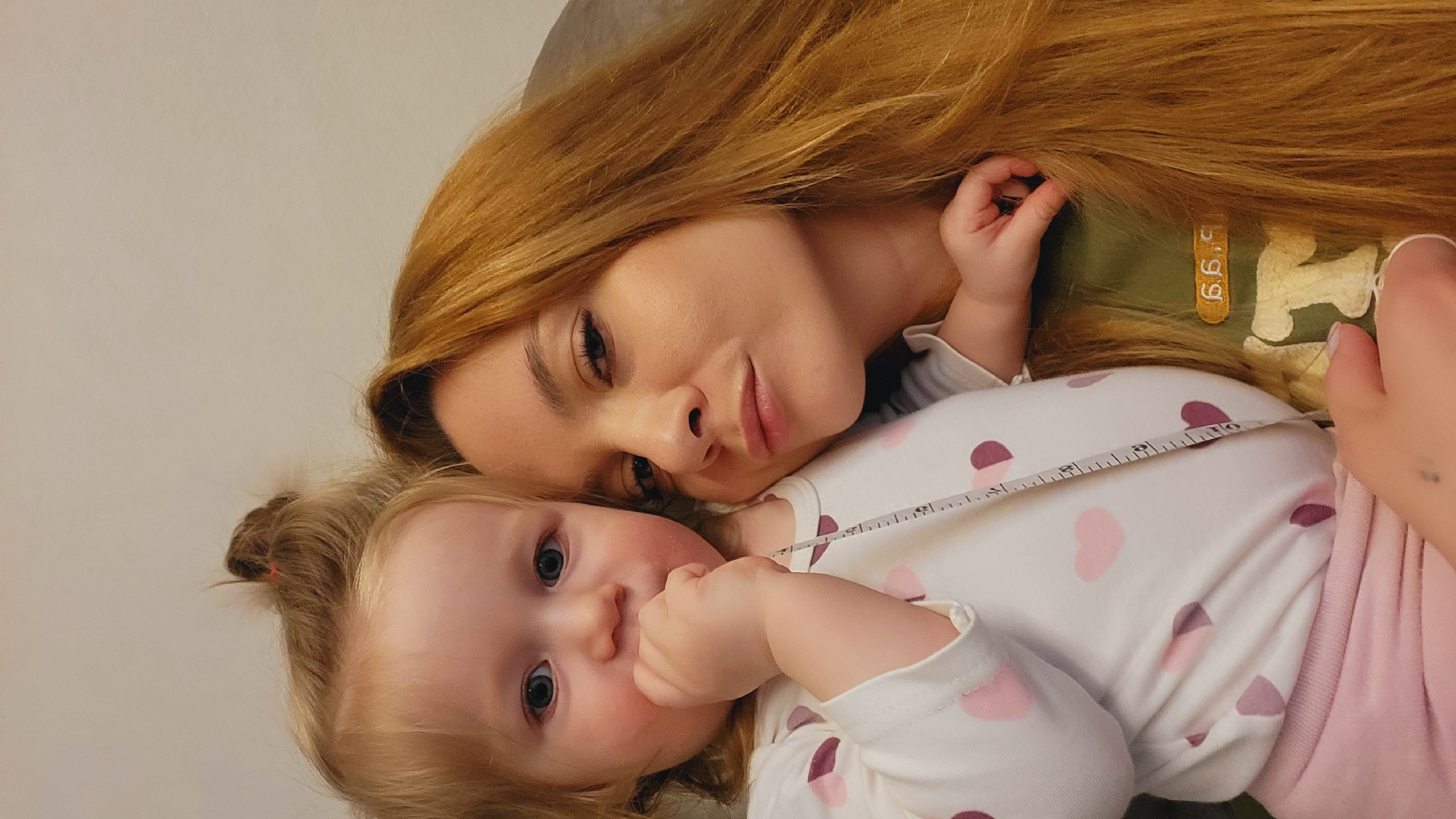 The image shows a woman and a young child lying close together. The woman has long, reddish-blonde hair and is wearing a green shirt. The child has blonde hair pulled back in a bun and is wearing a white shirt with pink and purple hearts. A measuring tape is draped across the child's stomach. The image appears to be taken indoors with a plain background. The location is not discernible from the image. There is no indication that the photograph was taken in Chaikovsky, Russia.