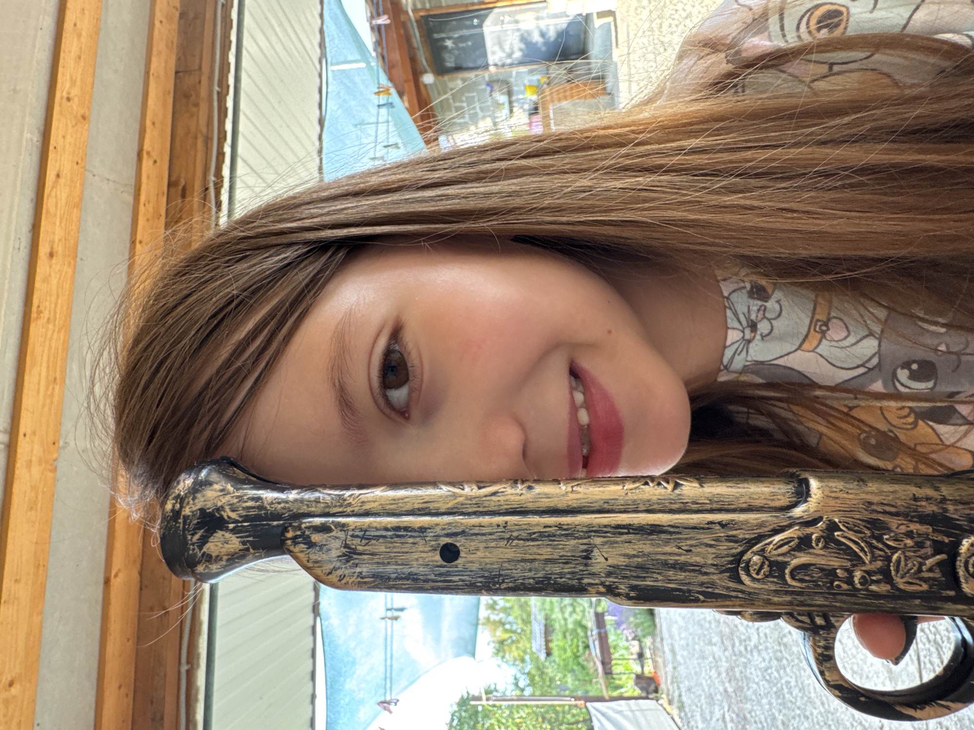 A young girl, with long brown hair and big brown eyes, smiles playfully into the camera, her head tilted and resting on a toy gun. Her lips are slightly parted, revealing her teeth, and her rosy cheeks suggest a youthful glow. She's wearing a light-colored shirt with a cute animal print. The toy gun appears to be a replica of an old-fashioned firearm, with a gold and black distressed finish and decorative etchings.
The background suggests an outdoor setting, possibly a yard or patio, with wooden structures and a glimpse of greenery and trees. The lighting indicates it's daytime, with bright sunlight filtering through, creating some glare and shadows. The overall mood is lighthearted and fun, with the girl's expression conveying joy and mischief. The image is shot vertically, making the scene appear as if it's viewed from the side. There is no visible text in the image. The location is Zassovskaya, Russia.