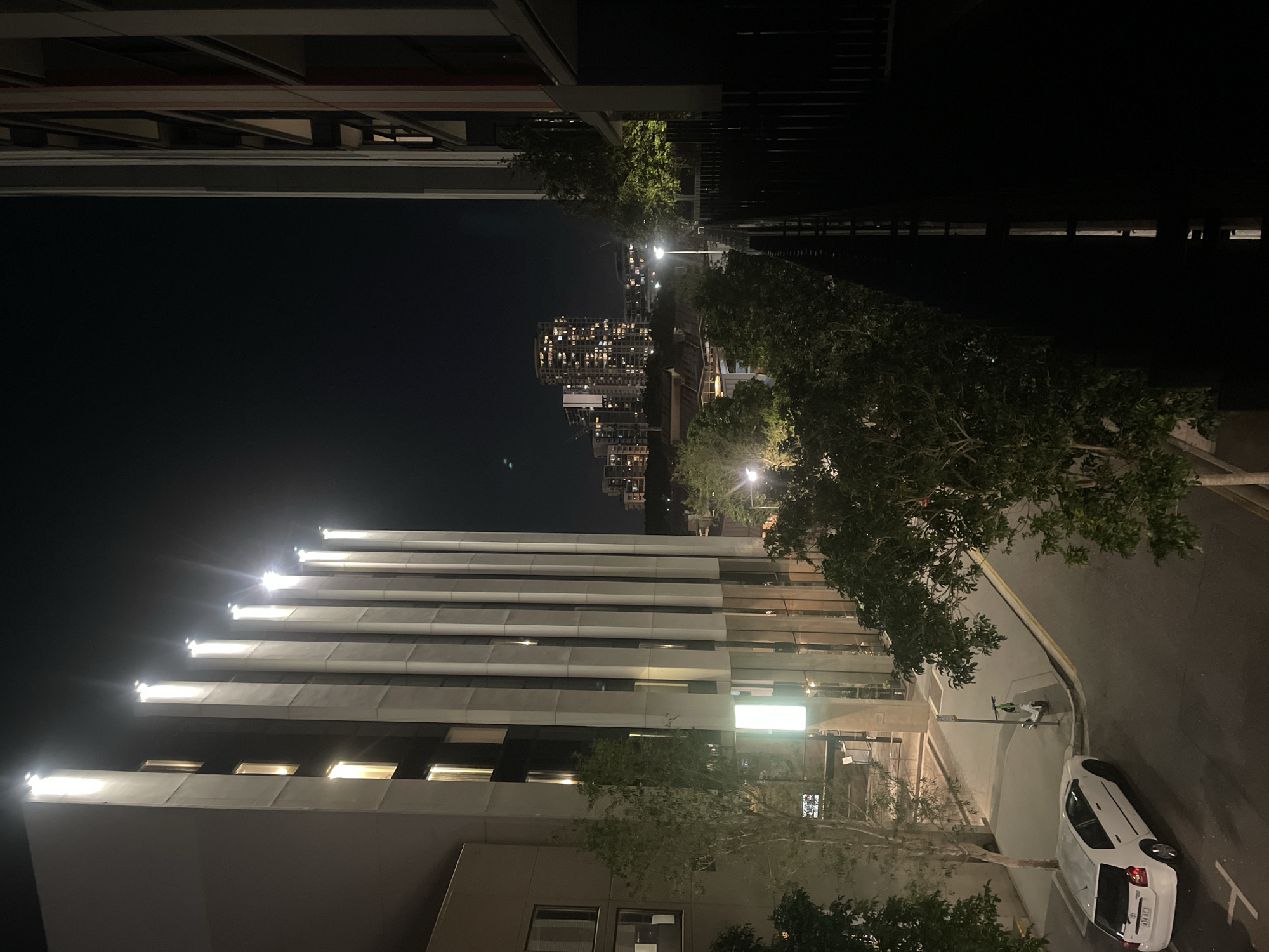 This image captures a nighttime view of a street in Brisbane, Australia, looking towards a cityscape. On the left, a modern building features a facade with vertical concrete fins, each topped with bright white lights that illuminate the night sky. Windows with warm interior lights are visible within the building. In front of this building, a white sedan is parked on the street. Trees with green foliage line the sidewalk and the street.
To the right, another building is partially visible, with balconies and a dark, enclosed lower section. The street itself is paved, with yellow and white lines marking its edges. Further in the distance, a dense cluster of high-rise buildings lights up the night, indicating the central business district of Brisbane. The overall atmosphere is urban and contemporary.
