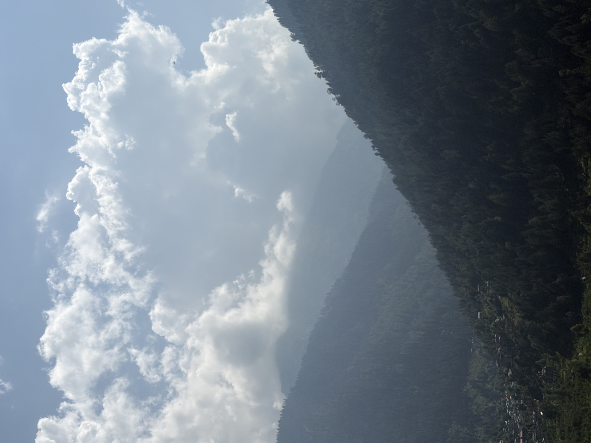 The image depicts a mountainous landscape in Vashisht, India, under a partly cloudy sky. The foreground is dominated by a steep, densely forested mountainside, with the trees appearing dark green and silhouetted against the lighter sky. In the mid-ground, a valley is visible, containing a small village or settlement with scattered buildings. Further in the background, more mountains rise, fading into a misty haze. Large, fluffy white clouds are prominent in the sky, casting some shadow on the landscape below. The overall impression is one of a remote, natural environment with human habitation nestled within the mountains. There are no people or specific actions clearly visible in the image.