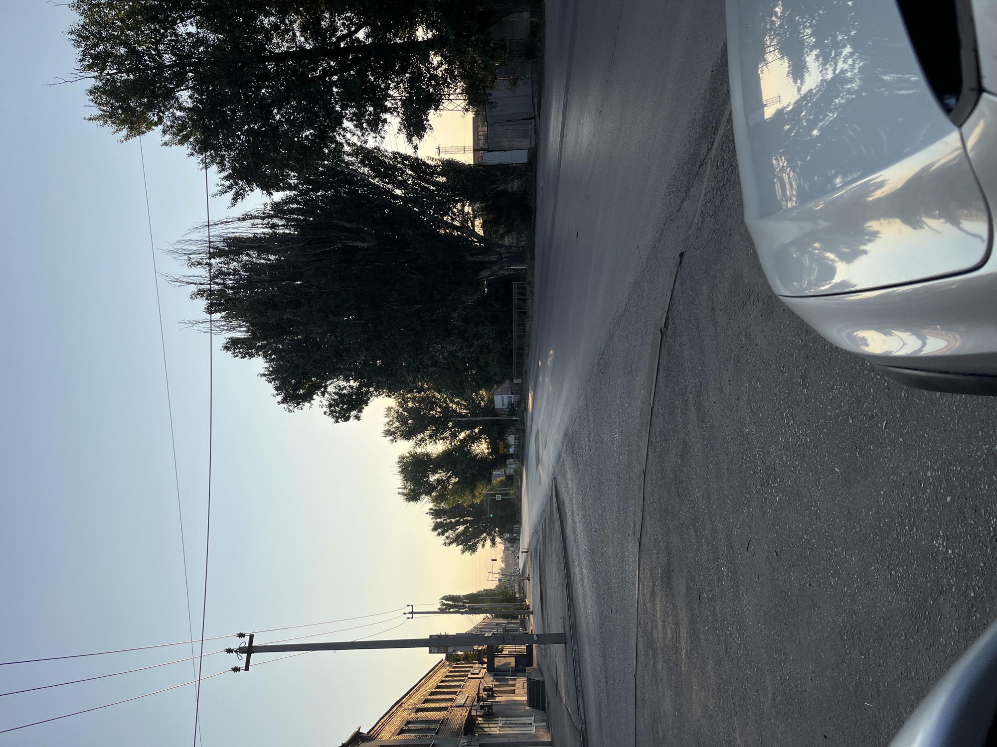 The image depicts a street scene in Taganrog City District, Russia, during the early morning or late evening, indicated by the low angle of the sun casting long shadows and the clear, pale sky. A paved road stretches into the distance, lined with trees on either side. On the left, a multi-story brick building with visible windows and doorways runs along the street. A utility pole with electrical wires stands prominently in the foreground, near the building. Further down the road, several more utility poles and trees are visible. The road surface appears somewhat wet or reflective, suggesting recent rain or dew. In the foreground, the hood of a light-colored vehicle is visible, reflecting the sky and surrounding trees. The overall impression is one of a quiet, ordinary urban street bathed in soft, natural light.