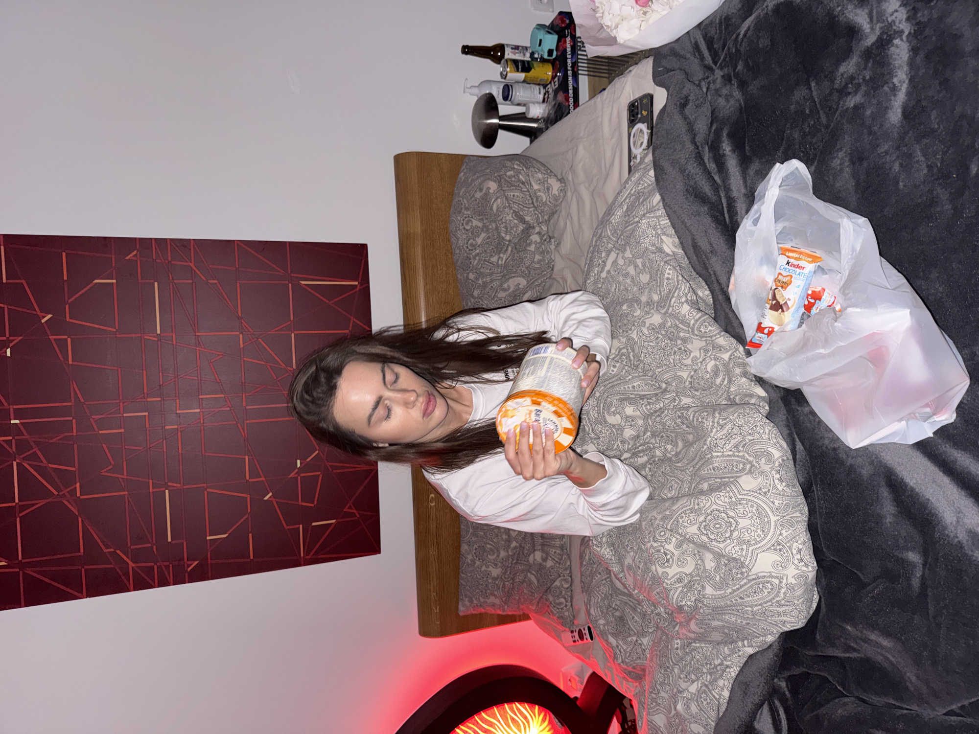 A young woman with long, dark hair lies in a bed in what appears to be a bedroom in Warsaw, Poland. She is wearing a white long-sleeved top and her eyes are cast downwards, suggesting relaxation or sleepiness. She holds a cylindrical container with an orange and white label in her hands.
The bed is covered with a grey and white paisley-patterned duvet and a dark grey plush blanket. Beside her on the blanket, a clear plastic bag contains a "Limited Edition Kinder Chocolate" bar. A smartphone with a patterned case rests on the bed near the headboard. On a bedside table, several bottles, a silver lamp, and a book titled "GOOD DESIGN IS FOR EVERYONE" are visible. The wall behind the bed features a large, dark red artwork with an abstract geometric pattern of lighter red lines. A device emitting a red, fiery glow is partially visible in the bottom left, providing ambient lighting to the scene. The setting suggests a cozy, personal space during the evening or night.