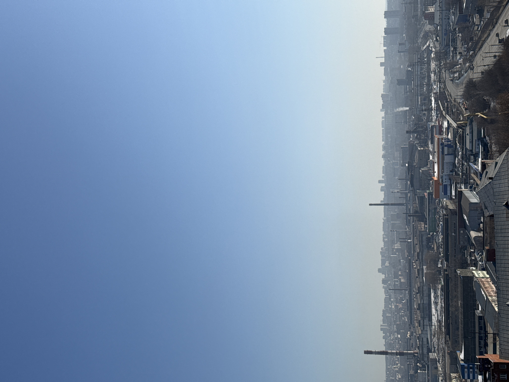 The image is a long-shot, high-angle view of a portion of the industrial area and city of Izhevsk, Russia. The scene is dominated by industrial structures including buildings with varying rooftops, pipelines, roads, and smokestacks extending into the sky. The sky has a blue gradient with lighter haze near the horizon and the city below.