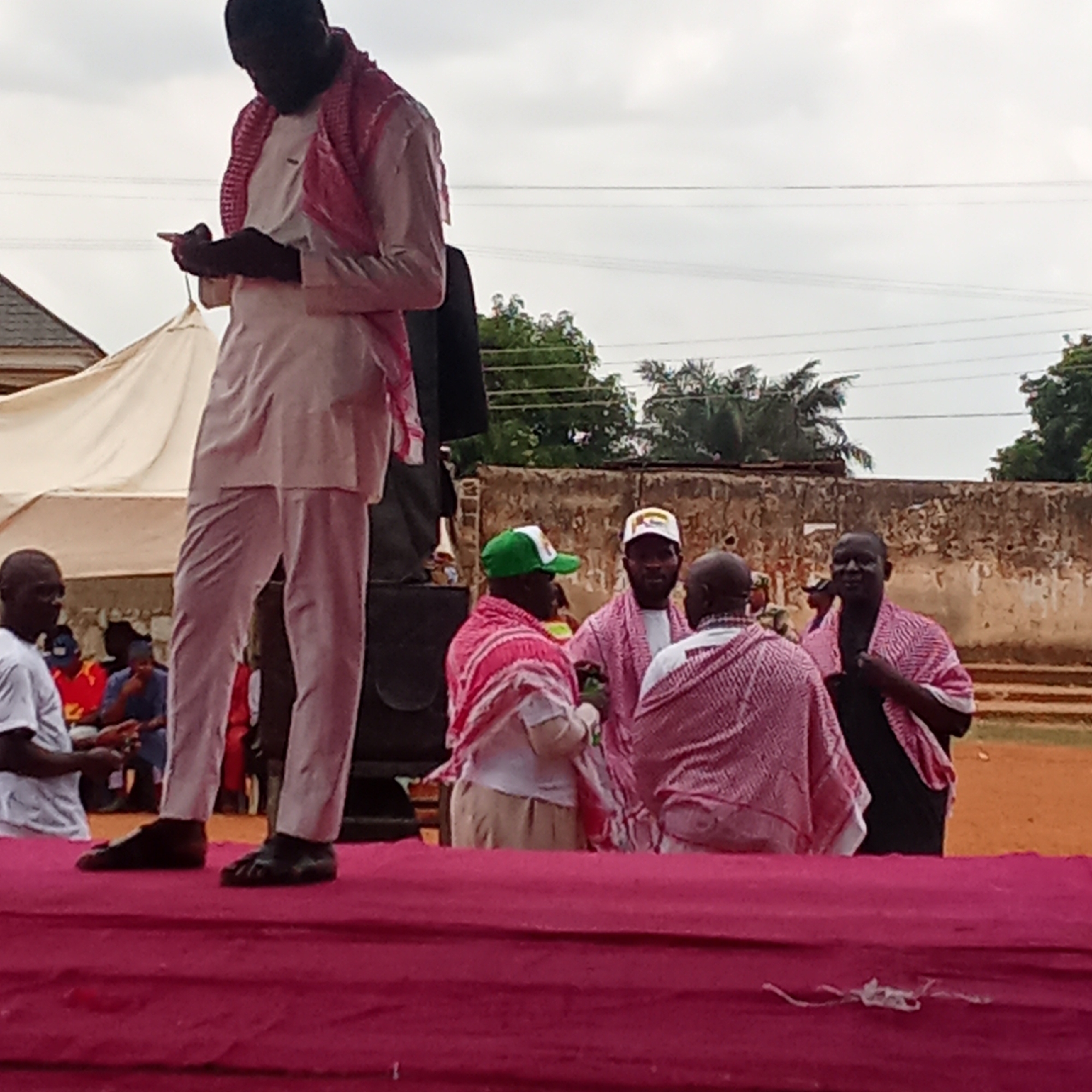 A photograph taken in Akwanga, Nigeria, shows several men on a raised red platform. One man, dressed in light-colored pants and shirt with a red and white patterned scarf, stands at the front of the platform looking down at something in his hands. Another man wearing a suit is standing behind a speaker. Three men standing closer together have the same scarf on. One wears a green hat, and another has a white hat. A few other people are seated and looking on. A white tent is visible behind the first man. Beyond the platform and men are a building and several trees. Overheard, powerlines stretch across the sky.