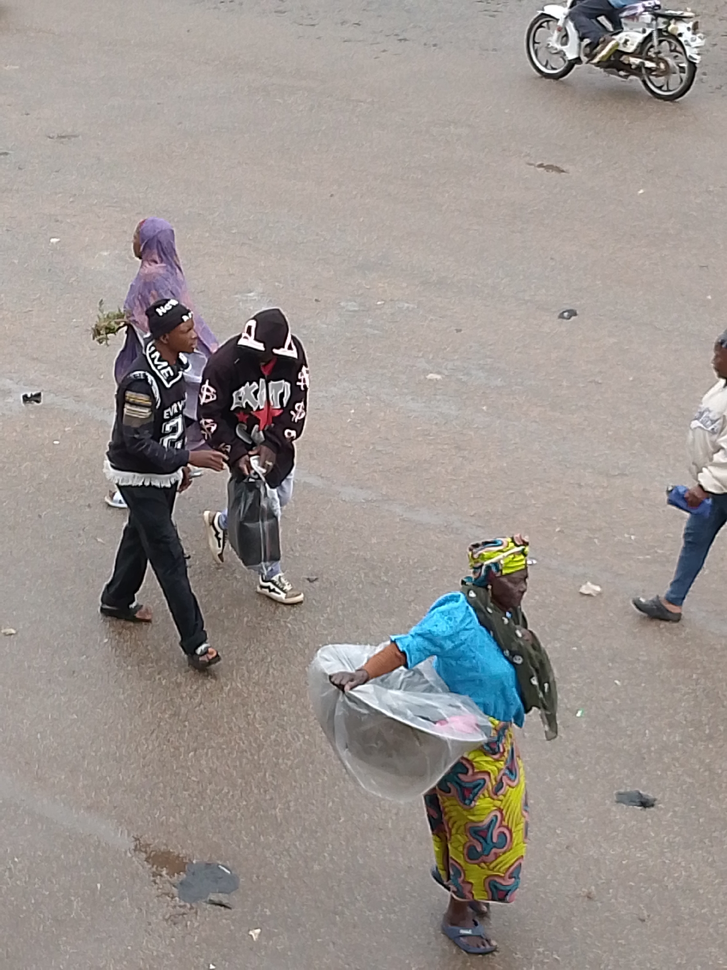 This high-angle shot captures a bustling street scene in Bukuru, Nigeria, during the daytime. The paved surface, possibly asphalt, shows some scattered debris.
In the foreground, on the right, a woman walks, dressed in a vibrant yellow, pink, and blue patterned long skirt, a bright blue top, and a dark green scarf. Her head is adorned with a colorful headwrap, and she wears blue flip-flops. She is carrying a large, round metal tray, covered with clear plastic, likely holding items for sale.
In the middle-left, two young men are walking together. The man on the left wears a black long-sleeved top with white lettering, partially visible as "SUPREME" and "EVERY," black pants, and sandals. He has a black beanie with "No" visible text. Beside him, the second man wears a black hooded sweatshirt featuring bold white and red designs, including what appears to be "AKIN" and dollar signs. He is carrying a black plastic bag and wears light-colored sneakers. Behind them, a person in a full purple garment, possibly a hijab or robe, walks while holding green leafy branches or flowers.
Further in the background, on the far right, another individual wearing a light-colored sweatshirt and blue jeans walks away, holding a blue object. At the top right, a person is seen riding a white motorcycle.
The scene depicts everyday activities, with people walking and a vendor carrying goods, suggesting a lively street environment.