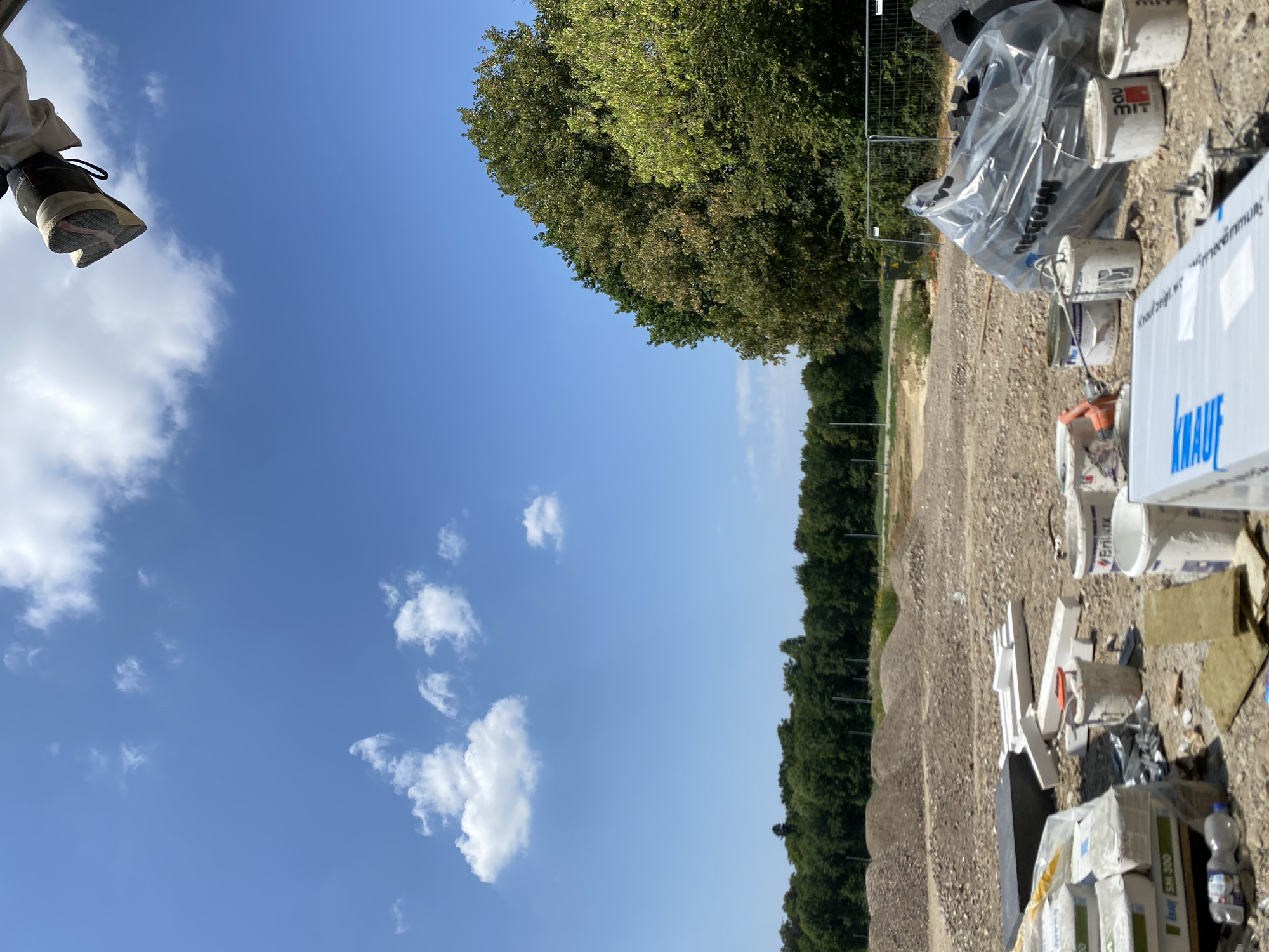 This image depicts a construction site under a clear blue sky with scattered white clouds. In the foreground, various construction materials are visible, including bags of what appears to be cement or plaster, white foam insulation pieces, and several buckets, some of which contain remnants of paint or plaster. A large, clear plastic-wrapped bundle of materials is also present. The ground is covered in gravel and dirt, with piles of excavated earth or rubble in the mid-ground. A line of lush green trees forms a backdrop, suggesting an outdoor, possibly rural or semi-rural, setting. In the upper right corner, a person's foot wearing a shoe is visible, seemingly resting on something above the frame, implying the photographer is positioned at an elevated viewpoint. The scene is indicative of ongoing construction work. The specific location in Hückelhoven, Germany, is not discernible from the visual elements alone, but the presence of construction suggests a developing area.