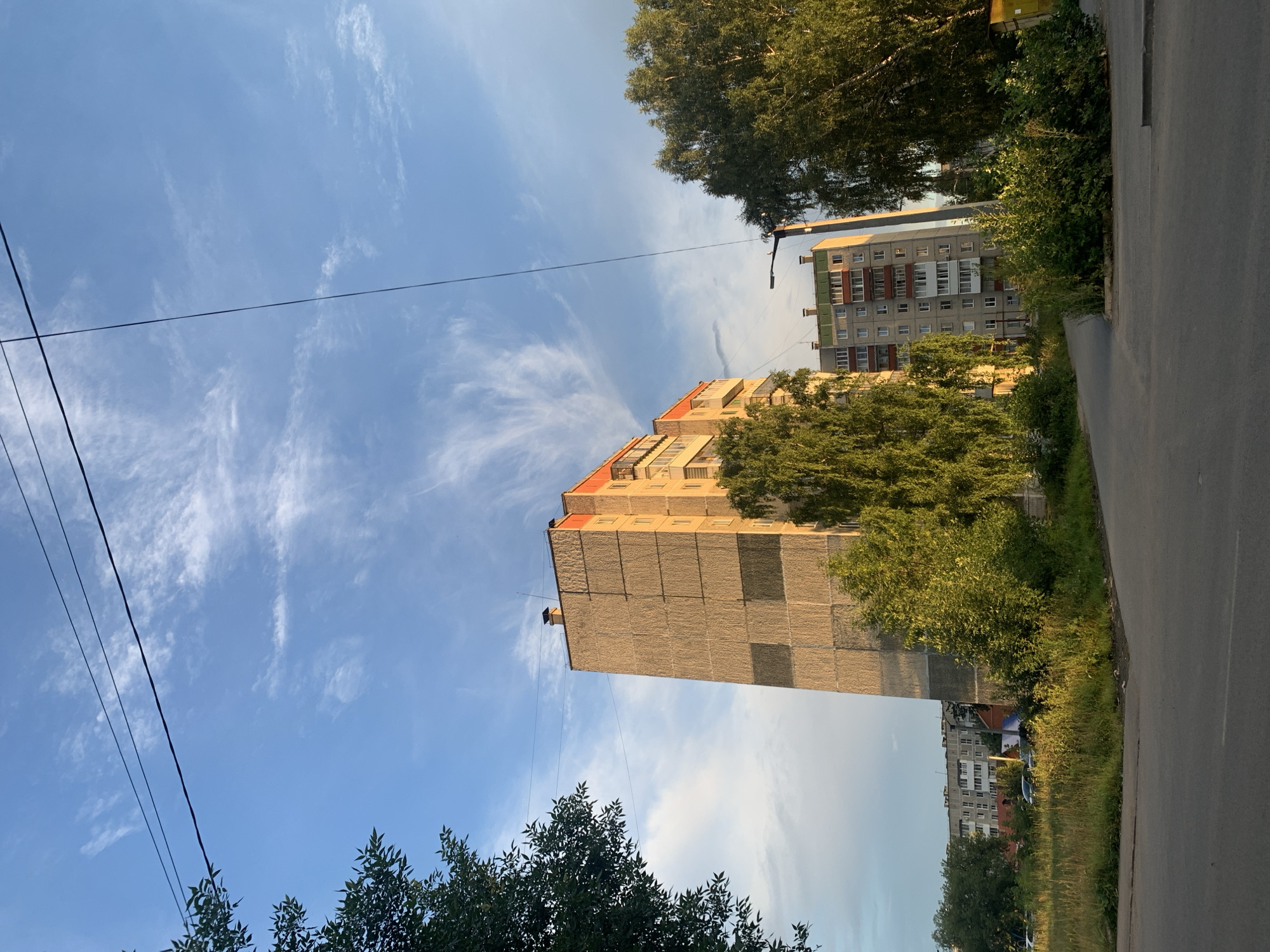 The image depicts a residential building in Asha, Russia, under a blue sky with wispy clouds. The building appears to be a multi-story apartment block constructed with concrete panels, some of which are discolored. The roofline features orange trim and chimneys. Trees with dense green foliage surround the building, with taller, mature trees on the right and left sides, and smaller trees and bushes closer to the structure. In the background, more apartment buildings are visible, along with a patch of overgrown grass and a paved road in the foreground. Power lines stretch across the sky, intersecting the scene. The lighting suggests it is either early morning or late afternoon, with warm sunlight illuminating the scene. No people are visible in the image.