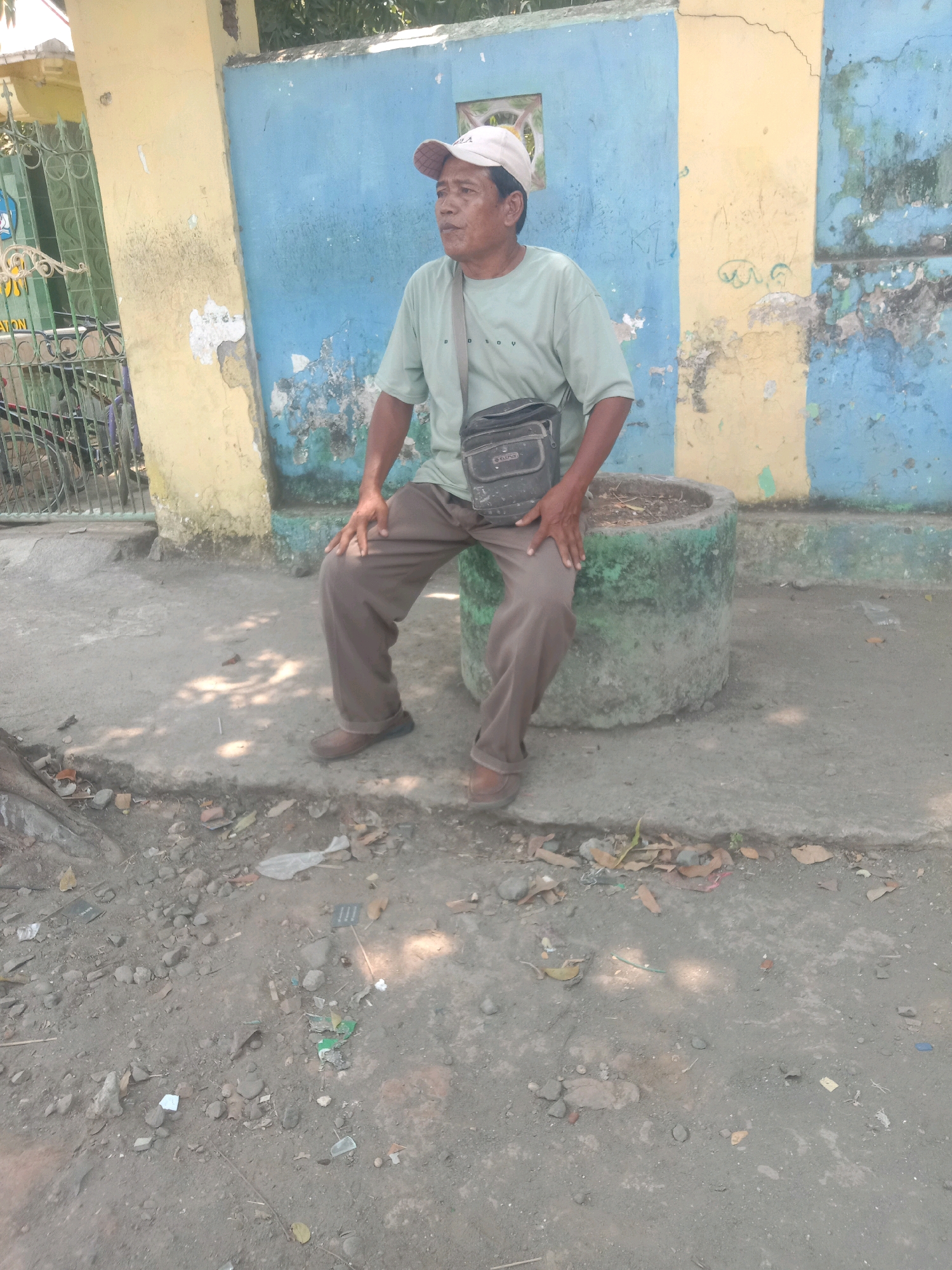 A man wearing a light green t-shirt, brown pants, and a beige baseball cap sits on a large, cylindrical concrete planter. He has a dark-colored satchel slung across his chest. The planter is situated on a grey, gritty surface that appears to be a sidewalk or unpaved area. Behind the man is a weathered wall painted in vertical stripes of yellow and blue, with sections of peeling paint and graffiti. To the left of the wall, a metal fence with decorative elements is visible, and behind it, a bicycle can be seen. The ground in the foreground is covered with dirt, small rocks, scattered leaves, and debris, including some pieces of plastic. The lighting suggests it is daytime, with shadows cast by the sunlight. This scene is located in the city of Ngempit, Indonesia.