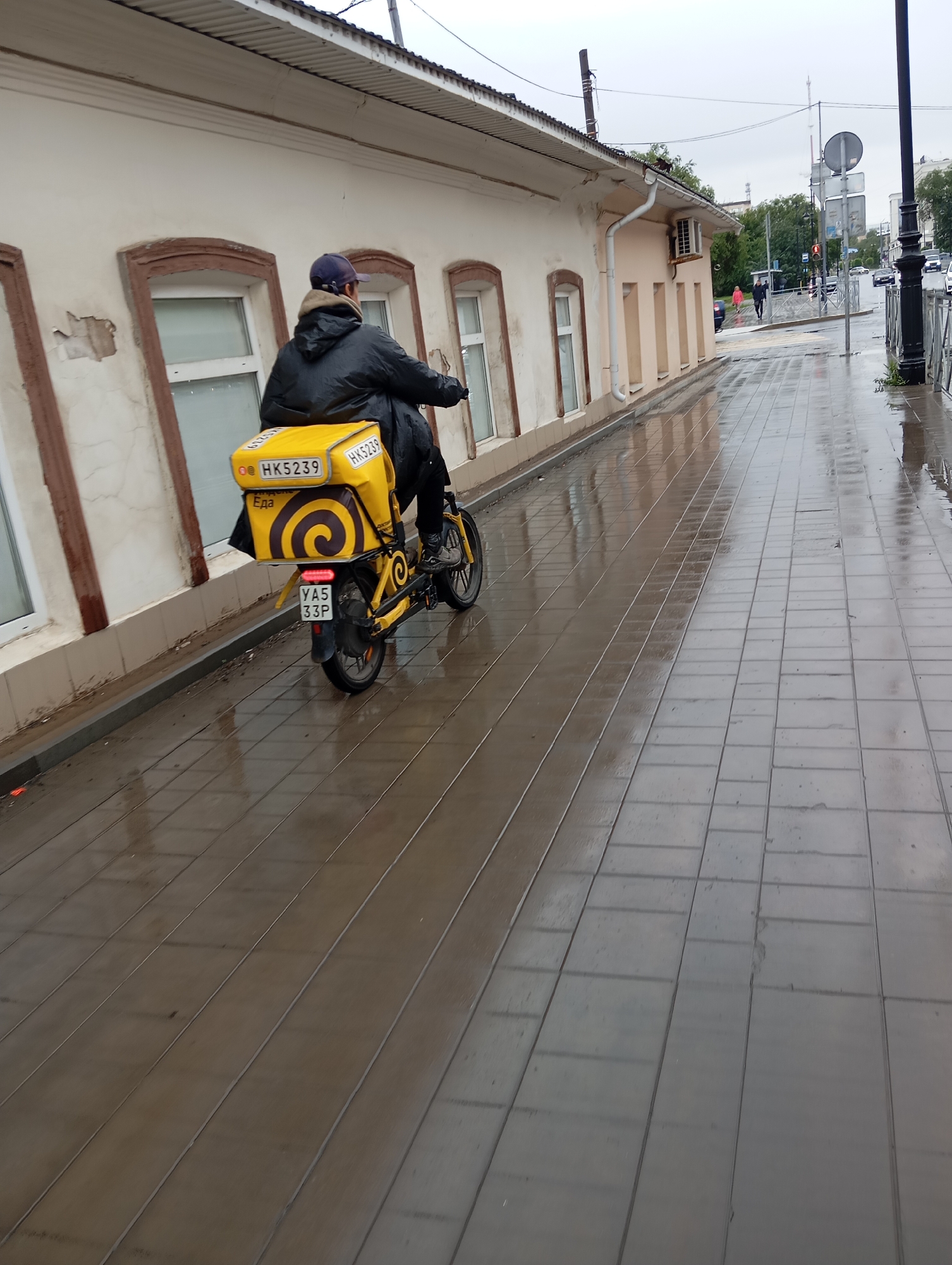 A person wearing a dark jacket and a blue baseball cap rides a yellow electric bicycle down a wet, paved sidewalk. The bicycle has a large yellow insulated delivery bag on the back with the logo "HK5239" and a spiral design. The license plate on the bicycle reads "YA5 33P". The person is viewed from behind and slightly to the side. The building to the left of the sidewalk is a light-colored, two-story structure with arched windows. The sidewalk is made of rectangular paving stones and reflects the overcast sky. In the background, a street with traffic and a pedestrian crossing can be seen, along with trees and other buildings. The location is identified as Orenburg, Russia.