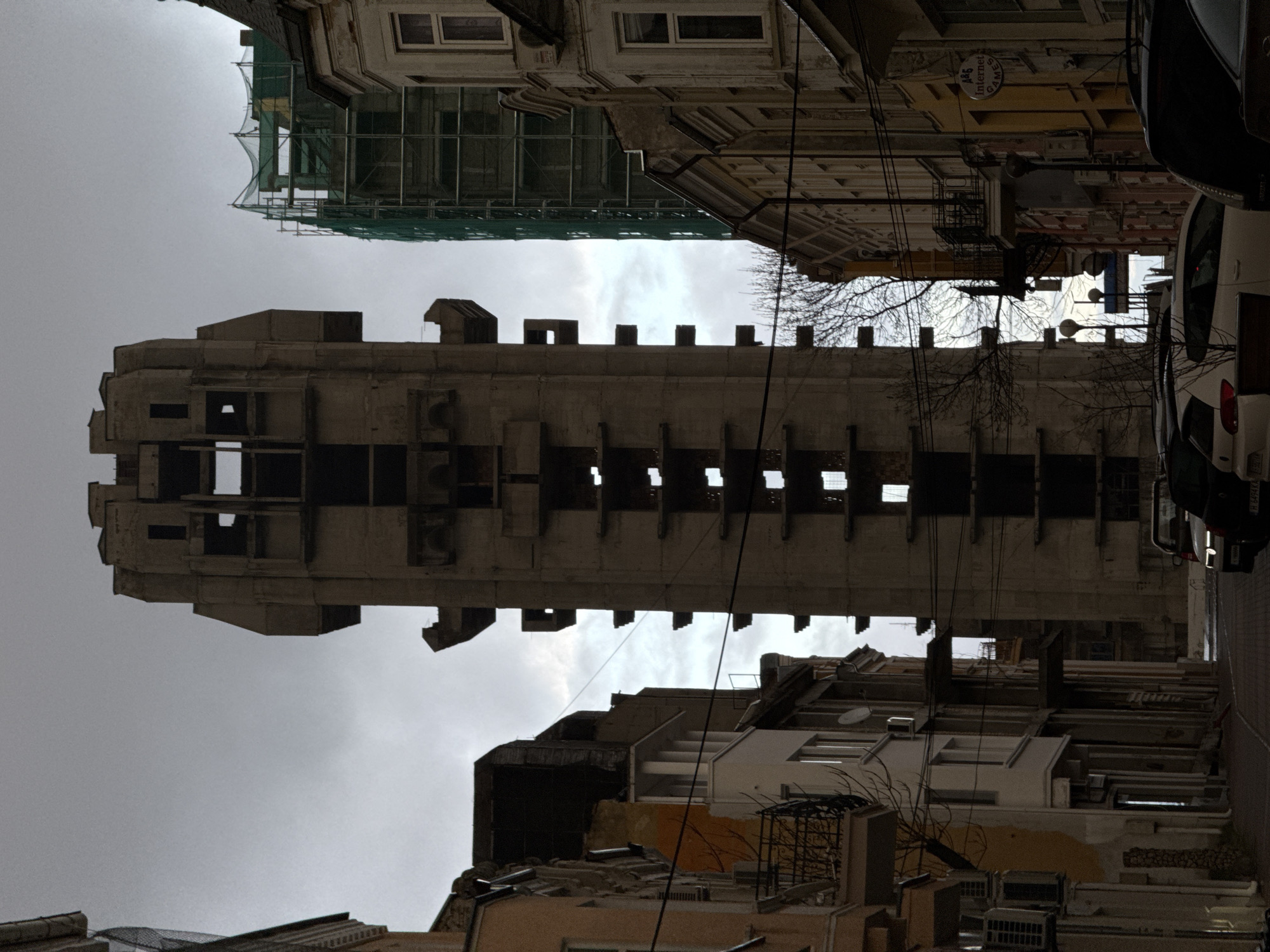 View looking up at the Central Post Office building in Shumen, Bulgaria. The sky is overcast. The Brutalist concrete building is in the center of the frame. Buildings are on either side of the Post Office, with scaffolding on the top left. The street to the right has parked cars. Power lines are visible between the buildings.
