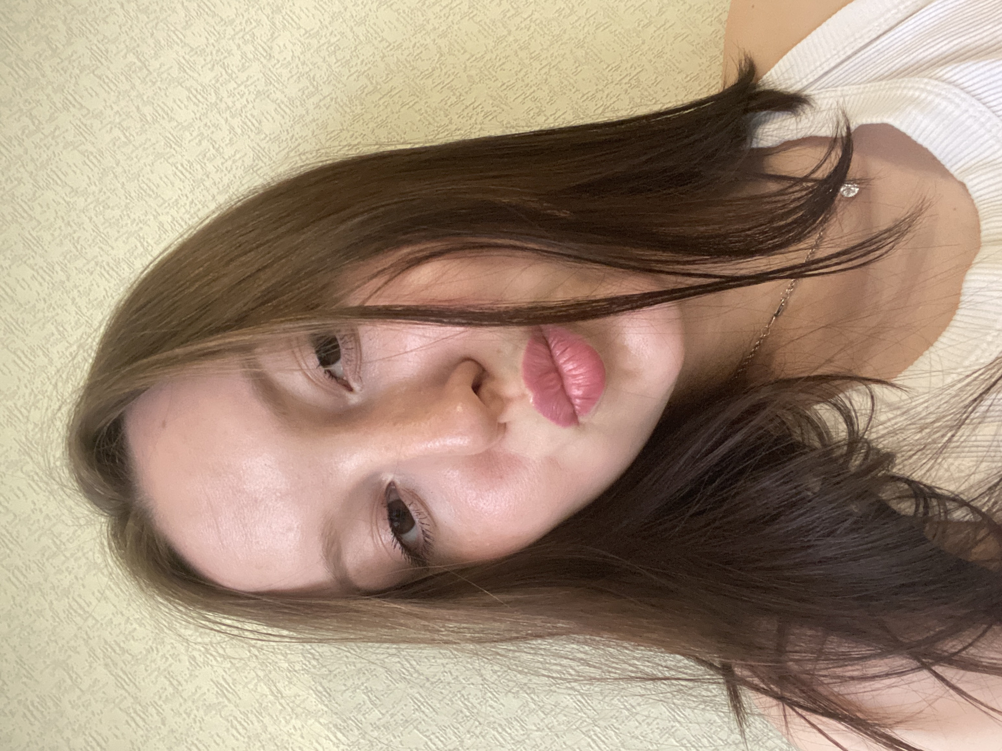 A young woman with long brown hair is positioned in the frame, her head tilted to the right. She is wearing a white top and a delicate gold chain with a small pendant. Her eyes are open, and her lips are slightly pursed. The background is a pale yellow wall with a textured pattern. The scene is likely indoors. The location is unknown and cannot be determined from the image.