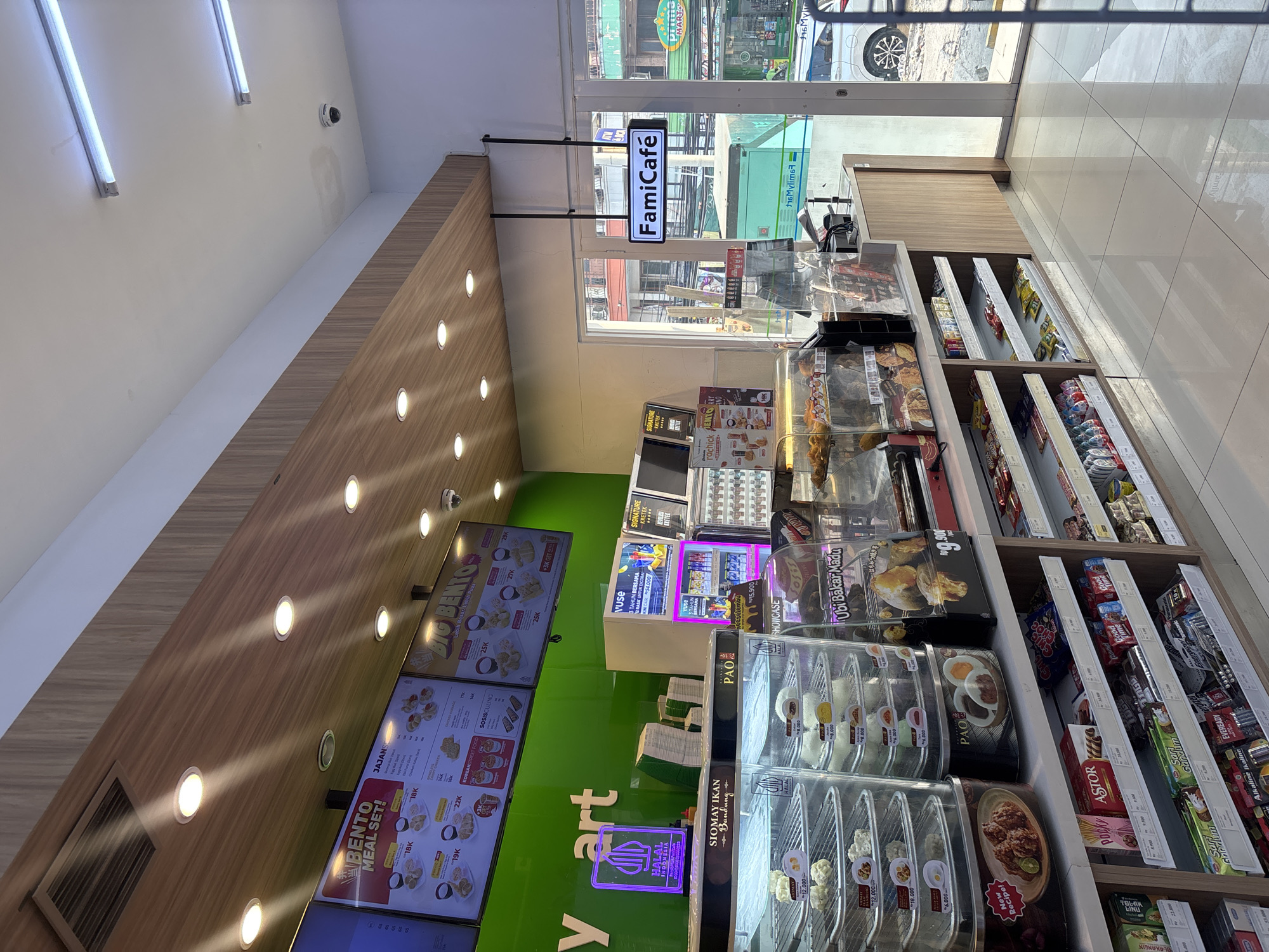 The image shows the interior of a brightly lit convenience store, likely a FamilyMart, in West Jakarta, Indonesia. The store features a green wall with a "Mart" logo, a wooden ceiling with embedded lights, and a variety of display shelves filled with food items and snacks.
In the foreground, there are refrigerated display cases filled with prepared foods like fried chicken and steamed buns. The prices are displayed in Indonesian Rupiah (Rp). Next to these, there are shelves stocked with packaged snacks, drinks, and batteries. Above the food displays, digital screens show menus and promotions.
A prominent sign hanging from the ceiling reads "FamiCafé". Through the glass doors in the background, a street scene is visible, with buildings, traffic, and a white car. The store appears clean and well-organized, with a focus on offering a range of ready-to-eat meals and convenience items. There are no people clearly visible in the image. The overall atmosphere is commercial and retail-oriented.