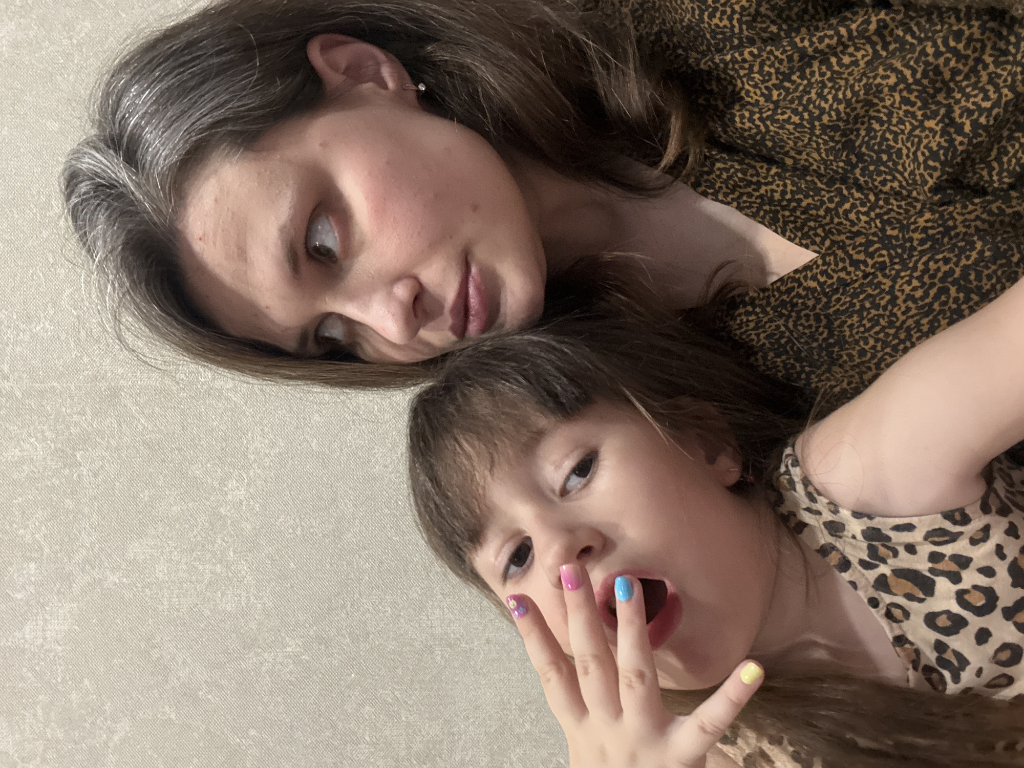 The image shows a woman and a young child, presumably a mother and daughter, lying down together. The woman is in the upper portion of the frame, looking down and to the side. She has light skin, brown hair with some graying strands, and is wearing a brown and black leopard-print patterned top. The child is beneath her, looking up at the camera with her mouth open and fingers raised, revealing various colored nail polish on her fingernails (purple, pink, blue, and yellow). She also wears a tan and brown leopard-print patterned shirt. The background is a neutral, textured surface, likely a wall or bed covering. The location is Chaikovsky, Russia, based on a reverse image search of the image.