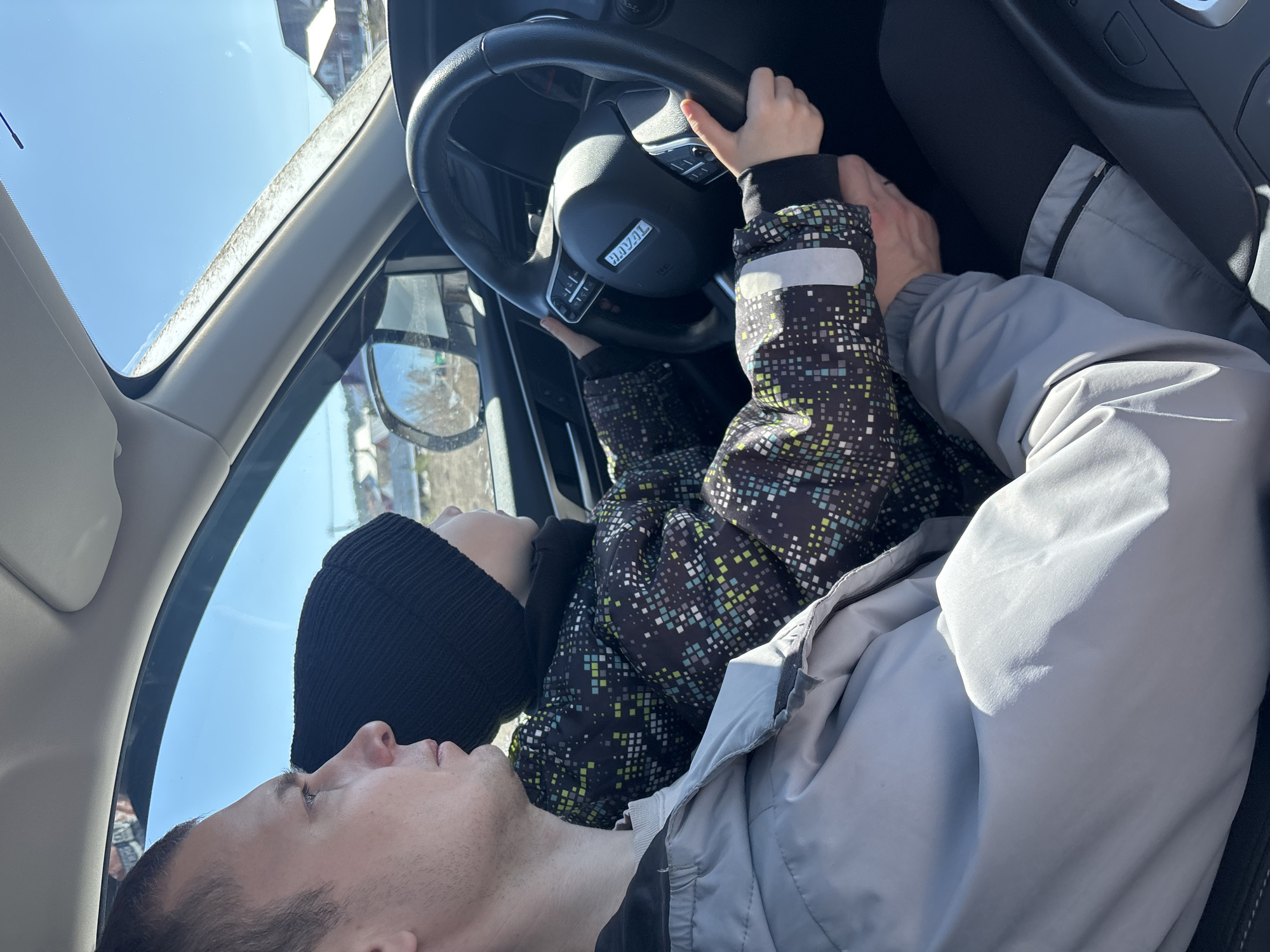 A man and a child are inside a car. The child, wearing a patterned jacket and black beanie, is reaching for the steering wheel, while the man, lying back with his head near the child, looks upward. The Haval brand steering wheel is visible, and the surrounding environment appears to be a street scene reflecting in the side mirror. The location is identified as the city of Chaikovsky, Russia.