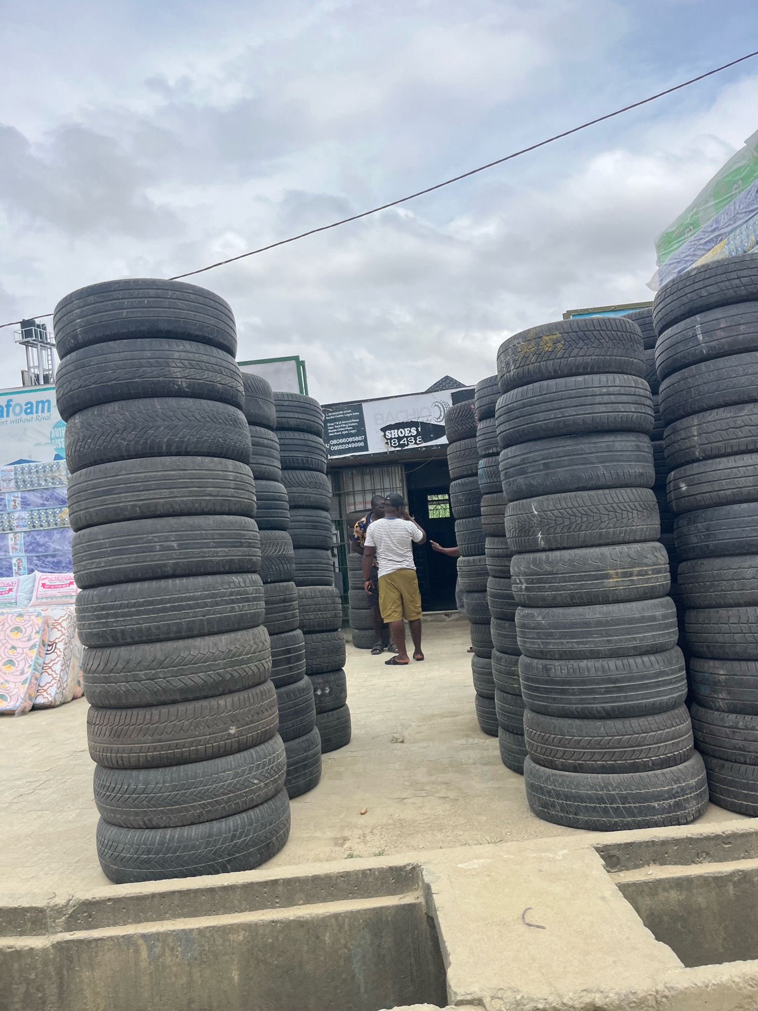 This image captures a scene in Ado, Nigeria, featuring a large outdoor storage area stacked with numerous tires. The tires are arranged in tall, vertical columns, with some columns reaching a significant height. In the background, there are buildings, including one with a sign that appears to read "BACHI SHOES" with a phone number. A few people are visible in the image. One man in a white shirt and shorts stands near the tire stacks, while another individual, wearing a striped shirt and shorts, is partially visible to the right. Another person is seen walking towards the background. The environment appears to be a commercial area with a slightly overcast sky. The ground is a paved surface, and in the foreground, there's a concrete structure that looks like a drainage channel or foundation. There are also stacks of mattresses or foam products visible on the left side of the image. The overall impression is one of a tire depot or a business involved in tire sales and storage.