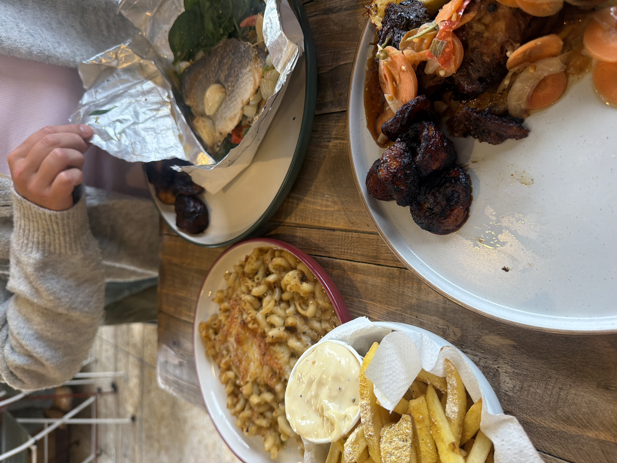 An overhead view captures a dining scene featuring multiple dishes on a rustic wooden table. One person is partially visible in the upper background, wearing a grey knitted cardigan over a light pink top. Their hands are engaged with a white, foil-wrapped package on a white plate with a dark green rim. This package contains a whole, cooked fish (identifiable by scales and shape), topped with several large garlic cloves and green leafy vegetables (possibly spinach), alongside visible red and white vegetable pieces within the foil. Next to the foil package on the same plate are several dark, roasted items, likely plantains.
To the left, a white bowl holds french fries, some dusted with a yellow seasoning powder. A small white paper napkin is nestled within the fries. A white cup containing a creamy, light yellow sauce with black speckles (suggesting seasoning) is placed among the fries.
Below the fries, a red-rimmed oval dish contains a baked macaroni dish with a golden-brown, slightly crusty top layer. The pasta appears to be elbow macaroni.
On the bottom right, a light grey speckled plate with a brown rim features dark, roasted or fried items (resembling plantains or meat), alongside a saucy preparation containing sliced orange carrots, white onions, and green elements, resting next to a portion of what appears to be grilled meat.
The background is blurred, showing light-colored interior elements, possibly furniture or architectural details, consistent with an indoor dining establishment. The image does not contain specific visual cues to identify the location as London, United Kingdom.
