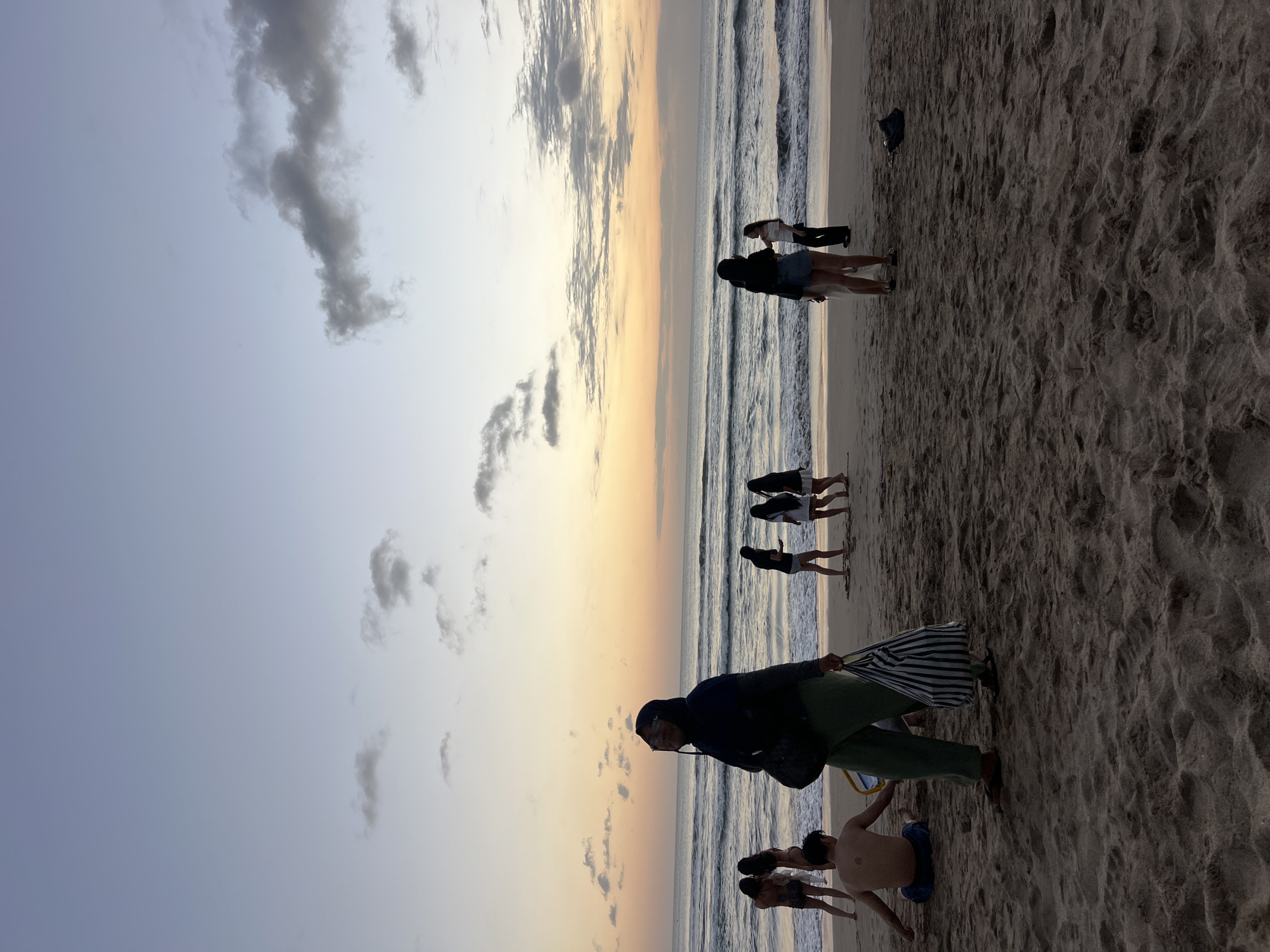 A vertical, wide shot captures a sunset on Kuta Beach, Indonesia, with the sky displaying a gradient of pale blue to soft orange. In the foreground, a sandy beach is dotted with several people. On the left, a person wearing a hijab and green pants walks towards the camera, carrying a striped tote bag. To their left and slightly behind, a shirtless person sits on the sand. In the mid-ground, a group of four people, three women and one man, stand near the water's edge. Two of the women are in swimwear, and one has their arm around the other. Further to the right, another group of three women are standing by the water. Two of them appear to be facing each other and talking, while the third woman stands slightly behind them. The ocean features moderate waves, and the horizon is marked by the distinct line of the sea meeting the sky. Several scattered clouds are visible in the sky, with the setting sun casting a warm glow. The sand is textured with footprints and ripples.