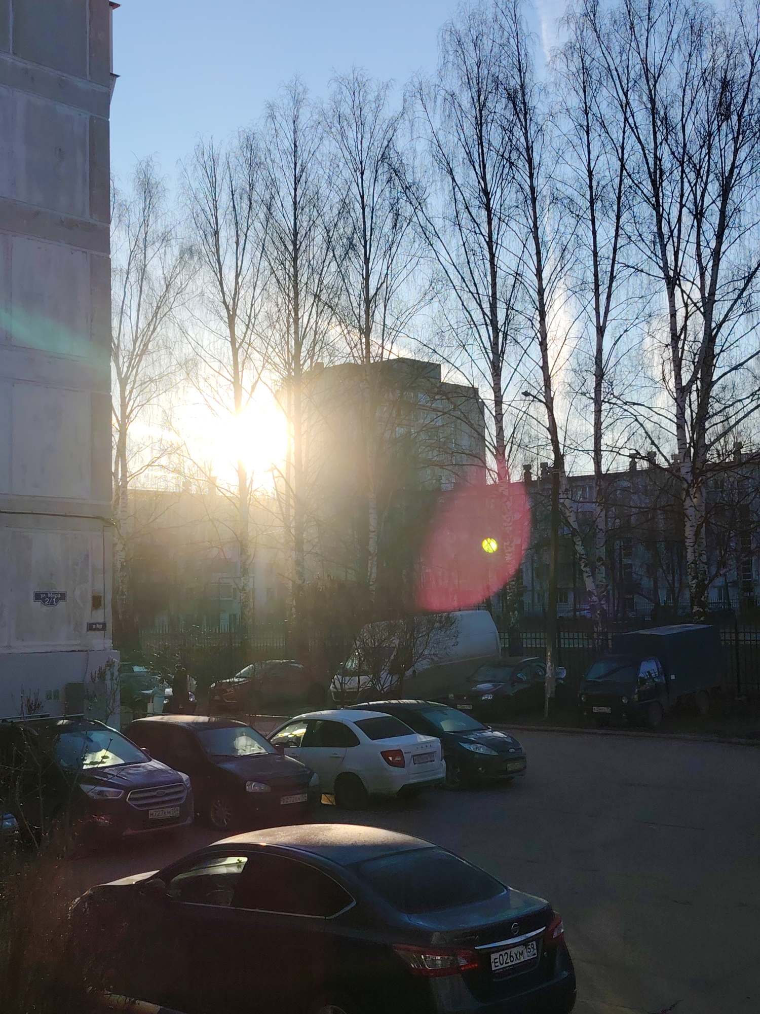 The photograph depicts a parking area in the city of Chaikovsky, Russia, at sunrise or sunset. In the foreground, several cars are parked. The nearest car, a dark sedan, has a license plate number E026XM 159. Other visible car models include a dark-colored Ford, a Lada, and a white car next to a green Ford.
Behind the cars, a low black metal fence separates the parking area from a building and trees. The sun is shining brightly, creating a lens flare effect. The trees are bare, suggesting winter or early spring. A multistory building can be seen between the trees. To the left is a concrete apartment building with a sign visible that reads, "Ya. Mupa 2/1".