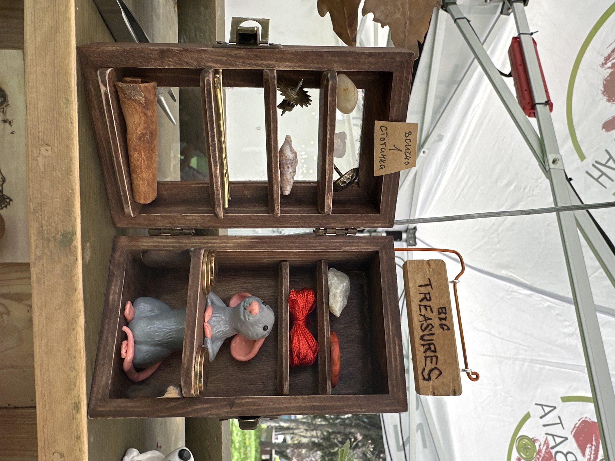 The image shows an open, wooden treasure chest with multiple compartments, each containing various small objects. The top section includes a piece of bark, a small gear, a shell, a rock, and a tag with text in Cyrillic, possibly indicating a price. The bottom section contains two gray clay mice, a small red bow, a rock, and a small wooden tag labeled "BIG TREASURES." The chest is positioned on a wooden table or stand, likely for sale. The setting appears to be an outdoor market or fair, with a white tent visible in the background. The location is potentially in Sofia, Bulgaria, based on the Cyrillic script on the price tag.