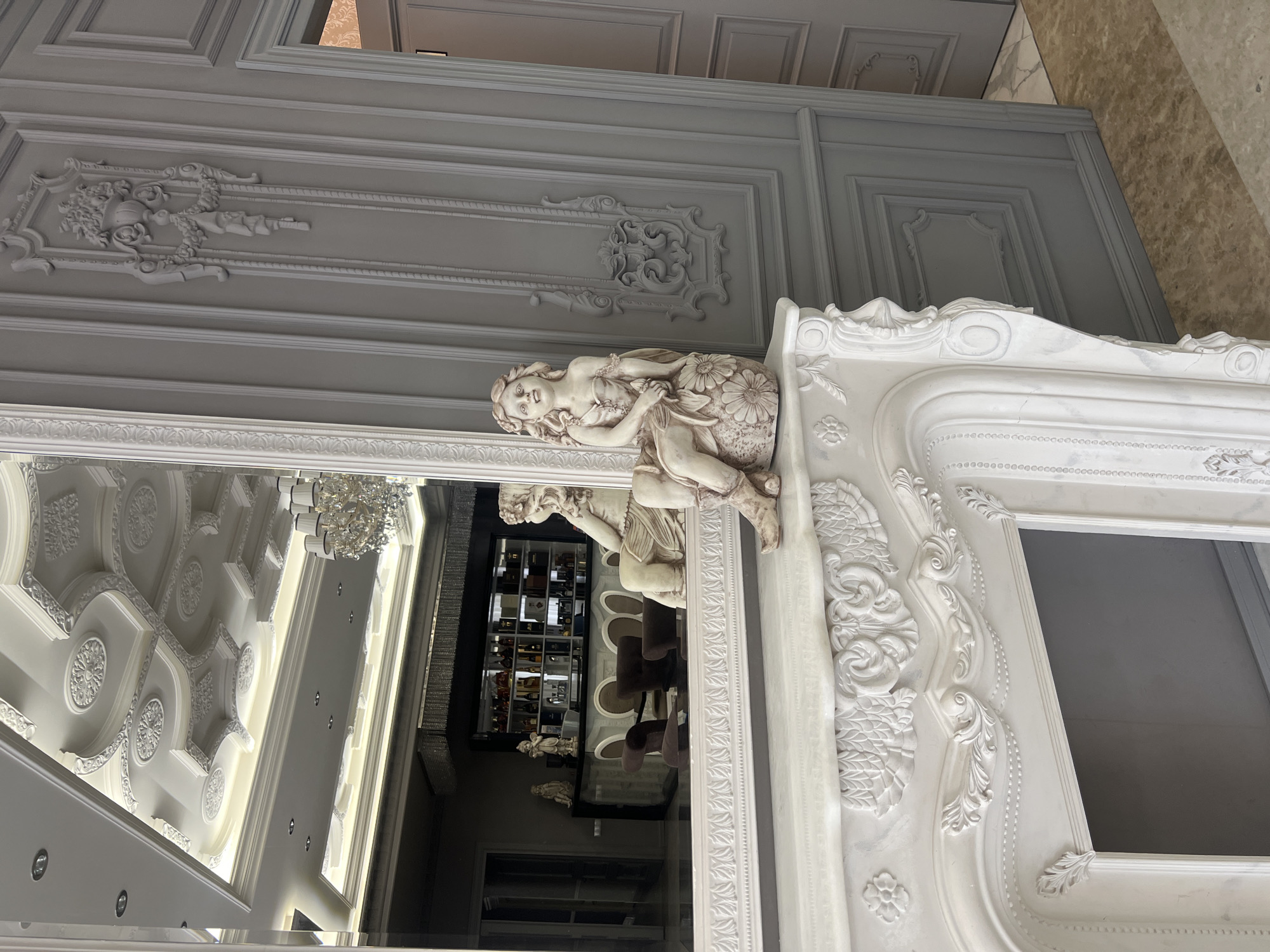 The image captures a highly ornate and luxurious interior space, dominated by elaborate classical design elements. In the foreground, a grand, light-colored (likely white or off-white) decorative structure, possibly a large mirror frame or a fireplace mantel, stands prominently. It is adorned with intricate carvings, including detailed scrollwork, floral motifs, and decorative borders. Perched on the upper part of this structure is a classical-style statue of a female figure, depicted sitting and holding or surrounded by flowers.
Behind this structure, a wall or ceiling paneling in a soft grey hue features elegant rectangular mouldings and ornate decorative embellishments, emphasizing a sophisticated aesthetic. A large mirror, occupying the left side of the frame, provides a reflection of another section of the interior. The reflection reveals a well-appointed bar area, complete with shelves stocked with numerous bottles and several bar stools. Above the bar in the reflection, an elaborate, multi-layered white ceiling with decorative mouldings, multiple chandeliers, and recessed lighting is visible, further highlighting the opulence of the setting.
The scene suggests a high-end commercial establishment or a grand private residence in Yerevan, Armenia, characterized by its detailed classical decor and lavish furnishings. The lighting indicates an indoor setting, but the time of day is not discernible. No specific activity is taking place, and no visible text is present.