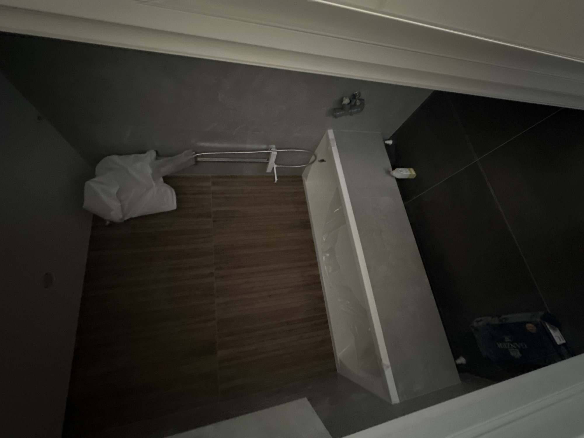 The image shows a bathroom under construction or recently completed. A white bathtub, still covered in plastic, is positioned against a wall with vertical wooden paneling. A showerhead and faucet are mounted on a grey tiled wall. On the floor, dark tiles are visible, with a bottle of cleaning product and a box with the label "GANZER" placed near the bathtub. A white doorframe is visible on the right side of the image. The scene appears to be located in Novoaltaysk, Russia, within a residential building. There are no people present in the image.