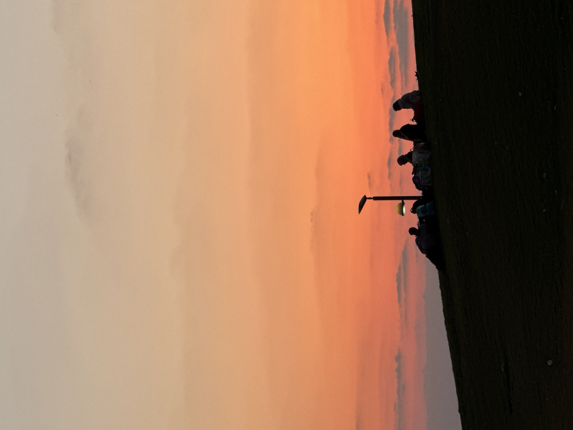 The image depicts a sunset scene on a grassy hill in Tel Aviv, Israel. Silhouetted against a vibrant orange and pink sky, a group of five people are gathered on the crest of the hill. They are seated and appear to be enjoying a picnic or a leisurely evening. A modern streetlamp with a distinctive curved design stands near the group. Several bags and a water bottle are visible among them, suggesting they are prepared for an extended stay. The foreground is dominated by the dark silhouette of the grassy slope, creating a stark contrast with the brightly colored sky. The overall impression is one of a peaceful gathering at dusk.