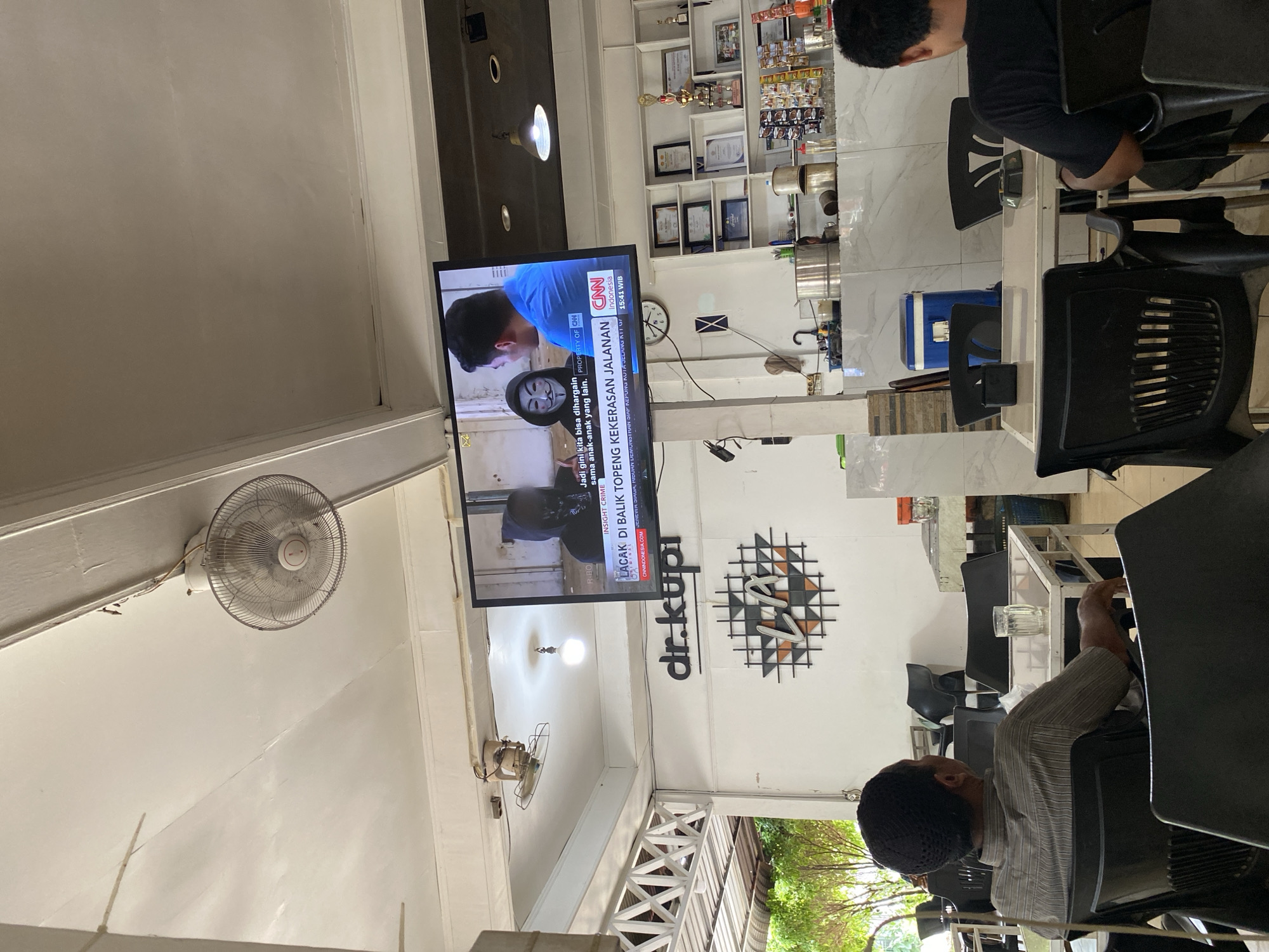 The image depicts the interior of a cafe in Lhokseumawe, Indonesia, identified by the "dr.kupi" signage on the wall. A television screen mounted on the ceiling displays a news report with the CNN Indonesia logo. The news report features an individual wearing a Guy Fawkes mask and another person speaking. A ceiling fan is visible in the upper portion of the image. The cafe environment includes tables and chairs, with patrons seated and facing away from the camera, observing the television. Shelving units behind the counter display various items, including framed certificates and trophies. The overall scene suggests a casual setting where patrons are engaged with the news broadcast.