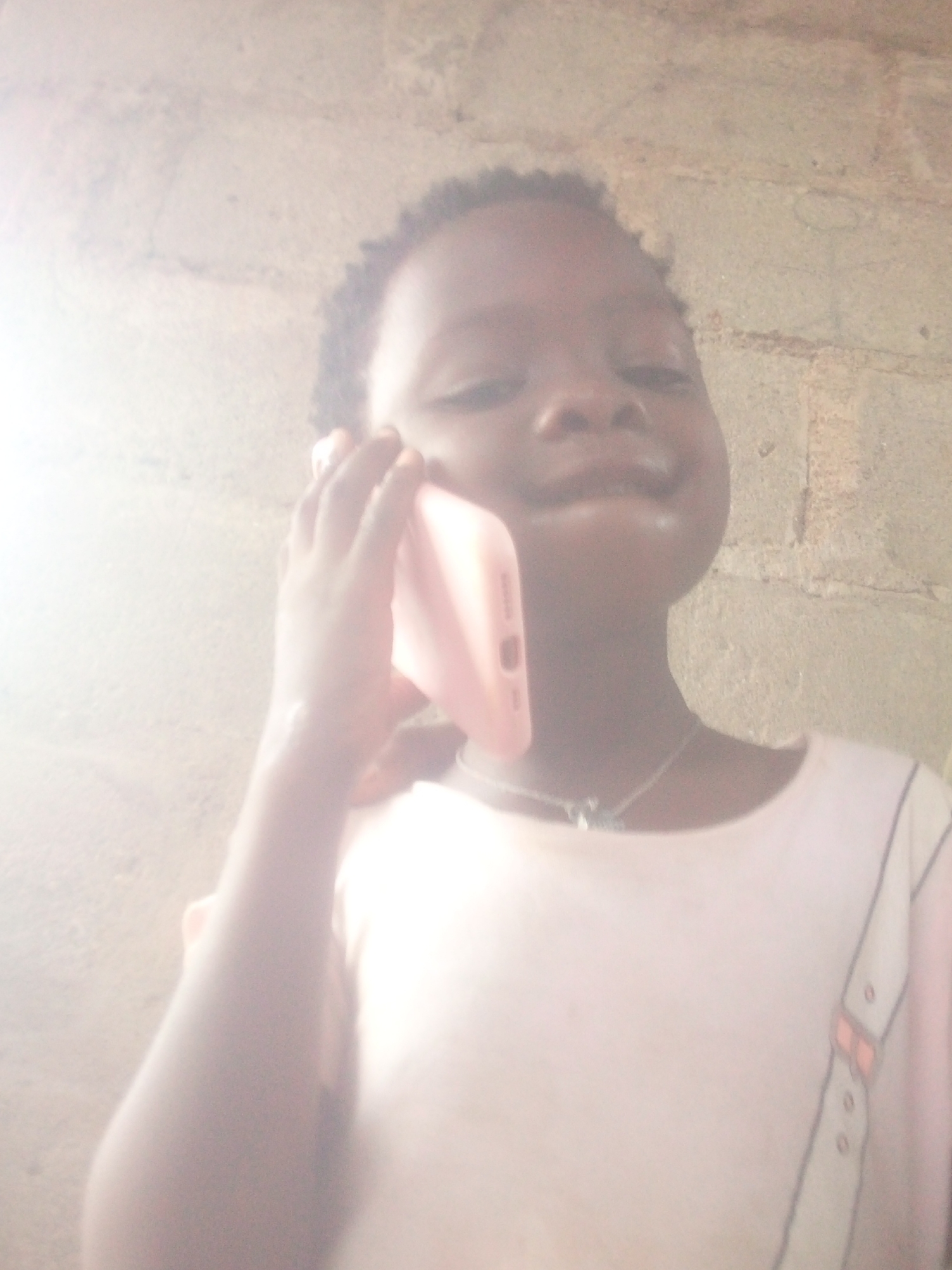 A young child, with dark skin and short, curly dark hair, is pictured from the chest up, holding a light pink smartphone to their right ear as if talking on a call. The child has a slight smile on their face and is looking towards the camera. Around their neck, a silver-colored necklace with a small, dark pendant is visible. They are wearing a light-colored, possibly white or very light pink, t-shirt that features a decorative pattern on the right side resembling a shirt placket or suspenders with outlined details and small, subtle orange or pink accents that look like bow ties or buttons.
The background consists of a light-colored, rough-textured wall, possibly made of concrete blocks, with visible horizontal and vertical lines suggesting mortar joints. The image is brightly lit, almost overexposed, particularly on the left side, indicating a strong light source, possibly natural daylight. The overall photo quality is somewhat soft and a bit blurry due to the bright conditions and possible camera limitations. There is no visible text in the image. The setting appears to be casual and possibly indoors or in a sheltered outdoor area.